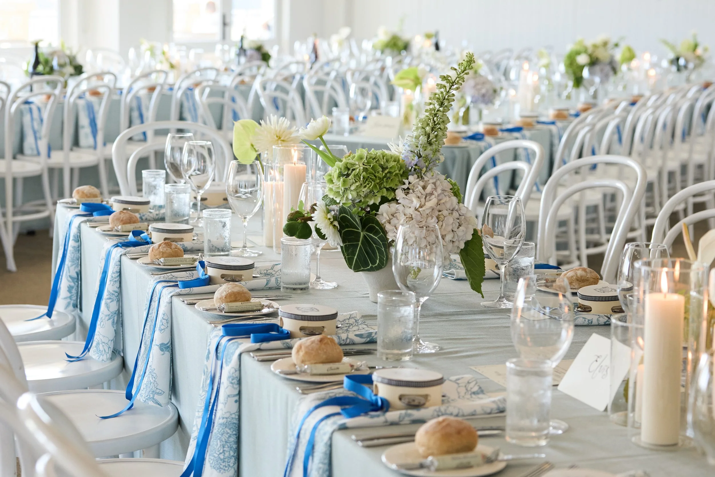 Elegant banquet table decorated with white and blue linens, floral centerpieces, candles, and place settings in a bright, airy venue.
