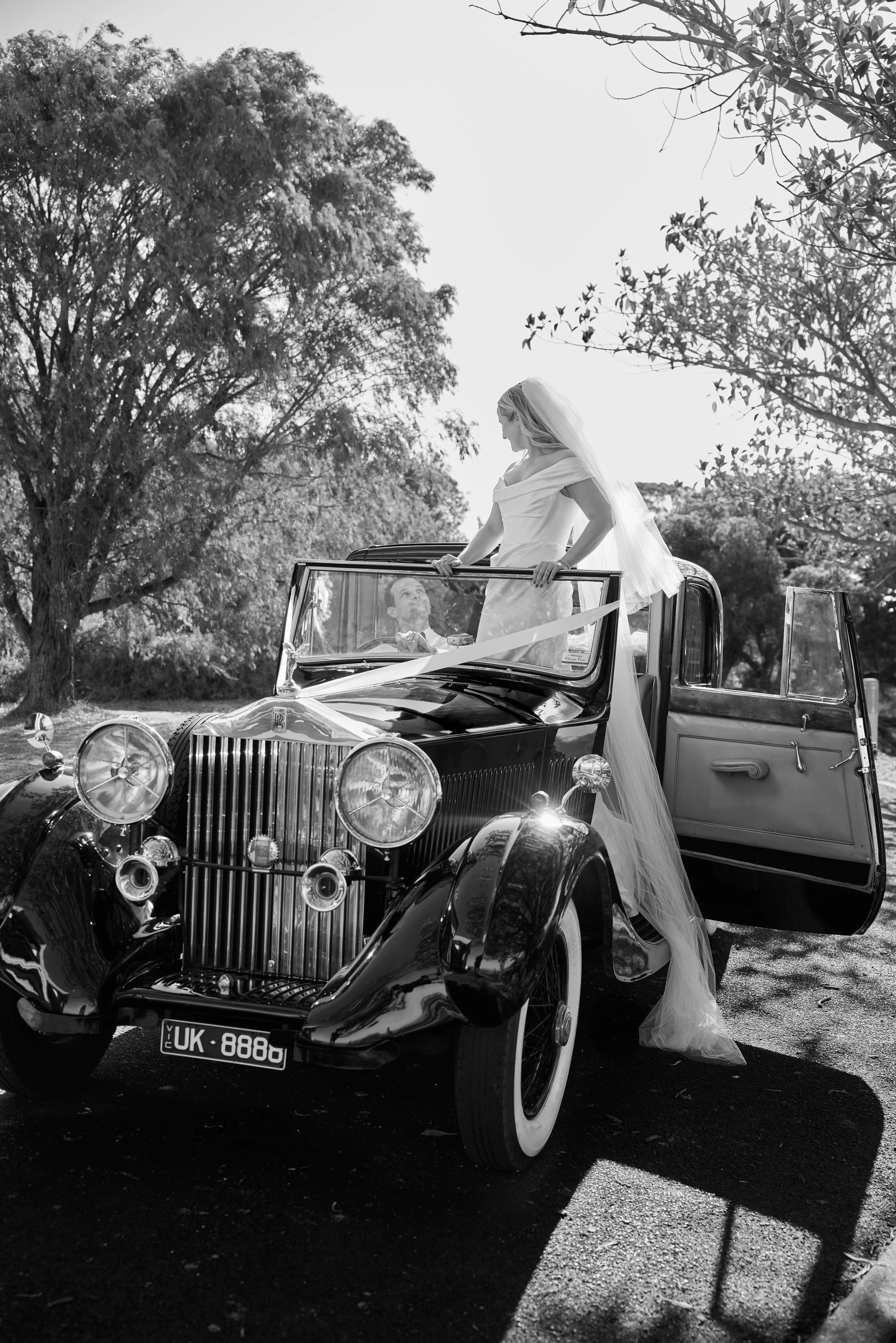 A bride in wedding dress standing on the hood of a vintage black car, looking down at a man sitting inside the car. The scene is outdoors with trees and sunlight filtering through the leaves.