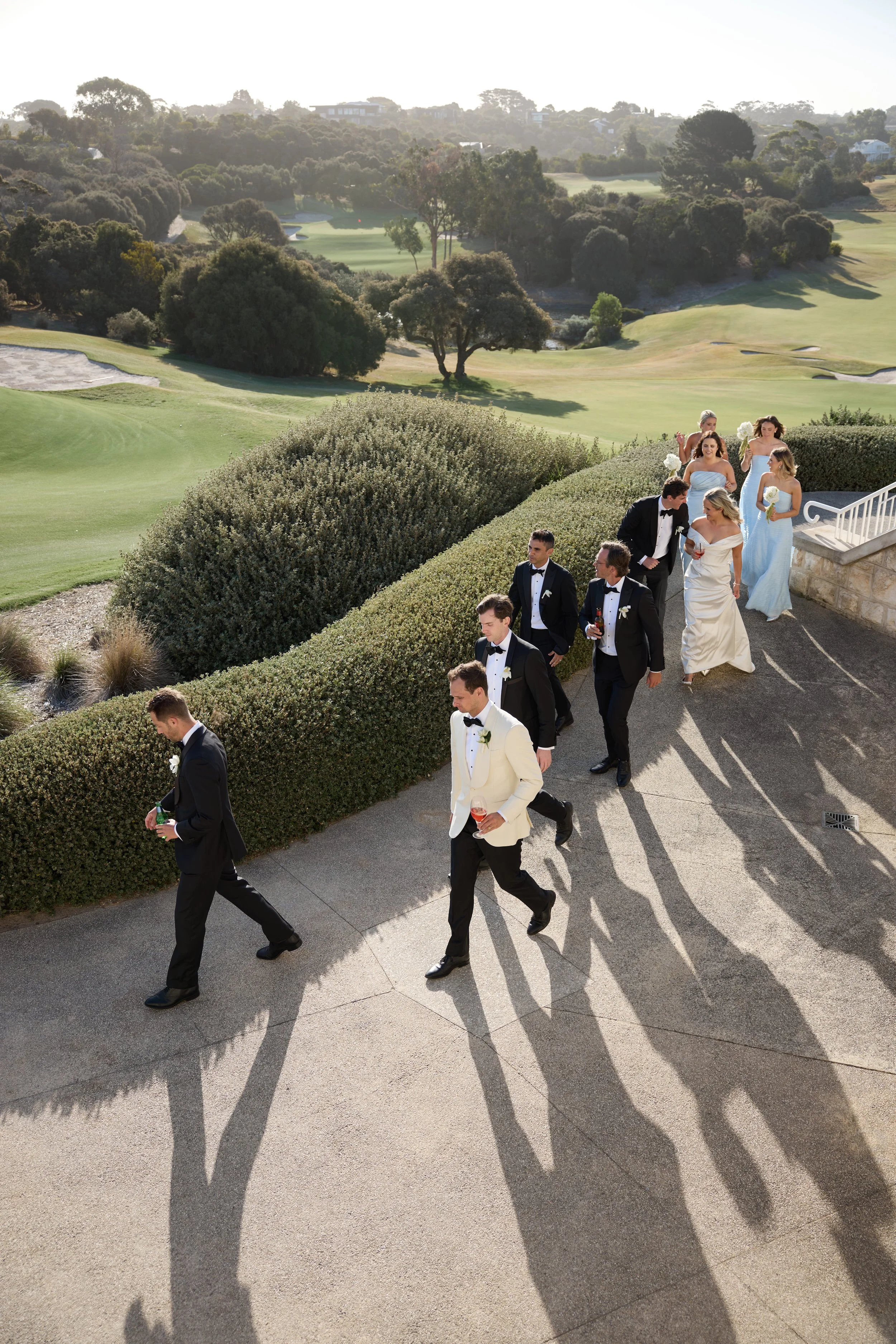 Group of wedding guests walking along a paved pathway at a golf course, with the sun casting long shadows and lush green trees and grass in the background.