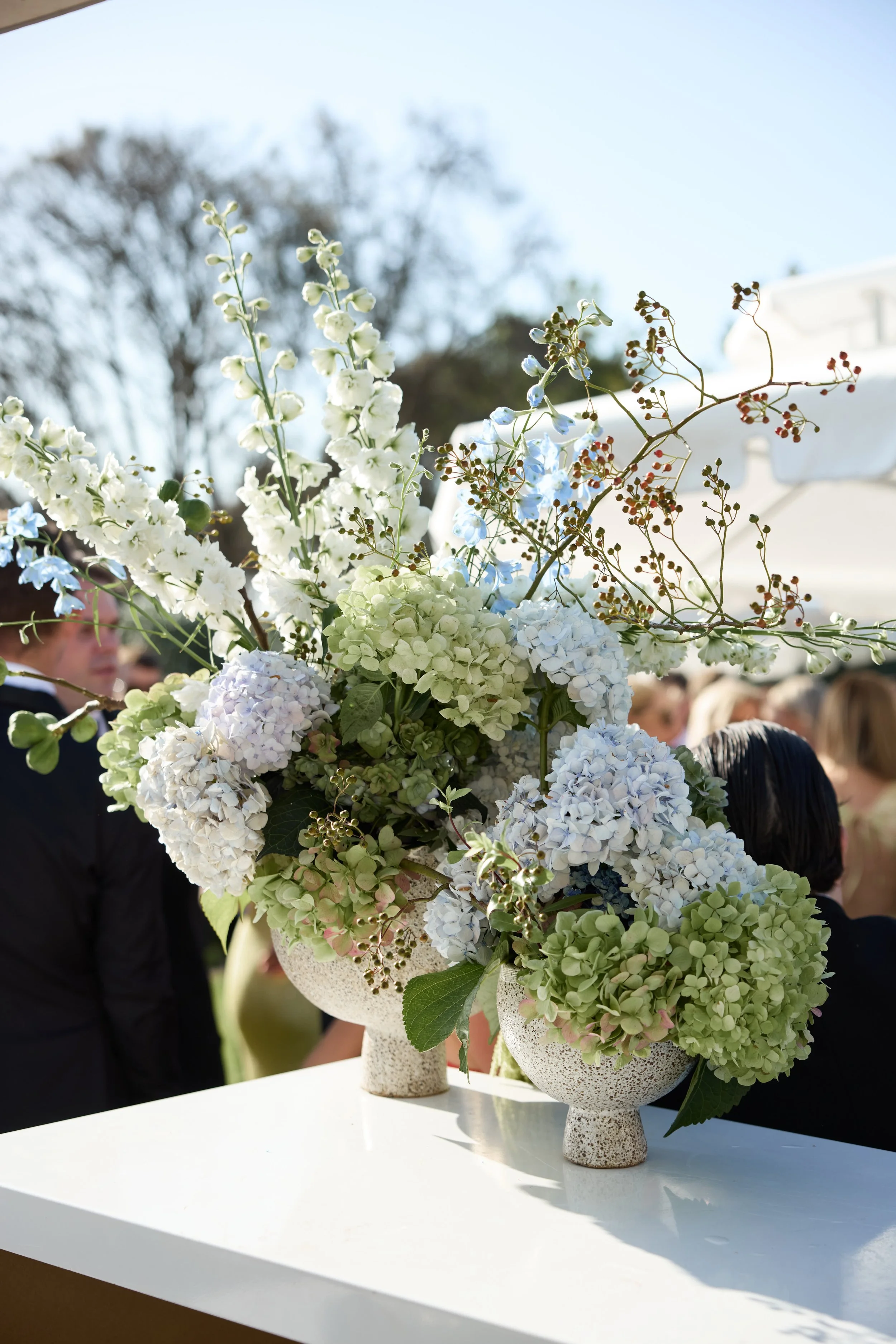 A large floral arrangement of white and pale green hydrangeas and other white flowers in two textured white vases on a white table at an outdoor event.