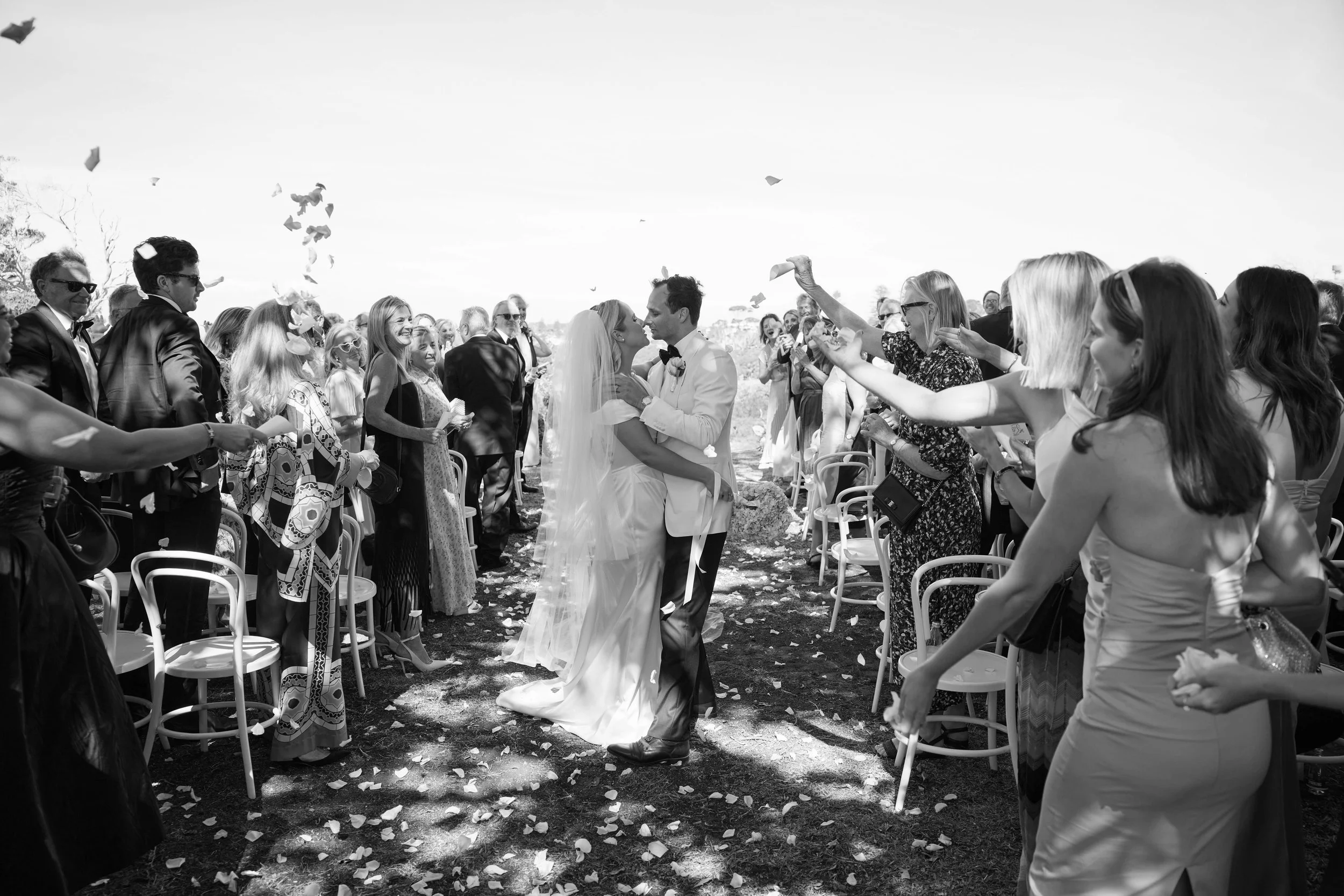 Black and white photo of a wedding scene with a bride and groom kissing while guests on both sides throw flower petals.