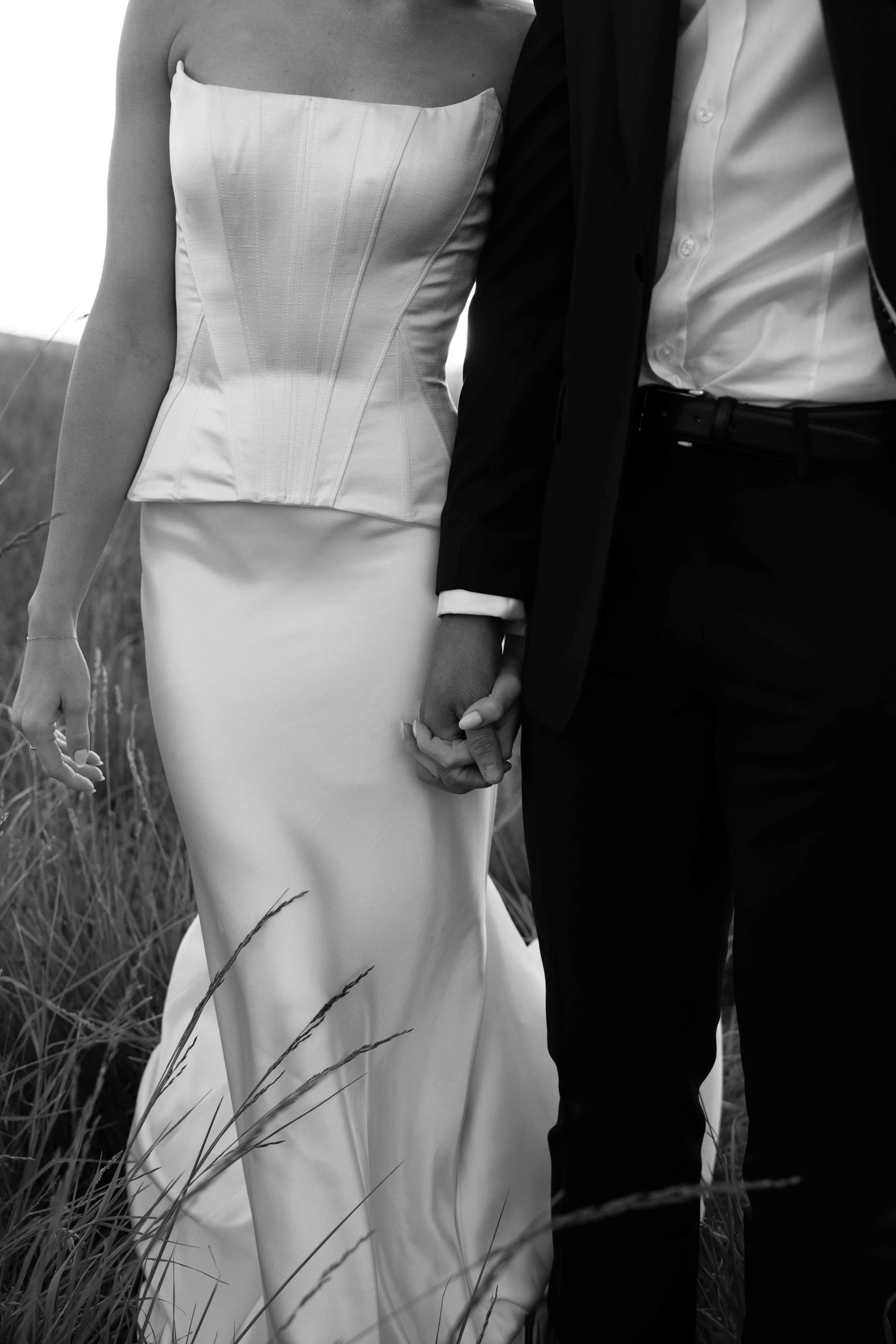 Couple dressed in wedding attire holding hands in a grassy outdoor setting. The woman is wearing a strapless white wedding dress, and the man is in a dark suit with an open-collared white shirt.