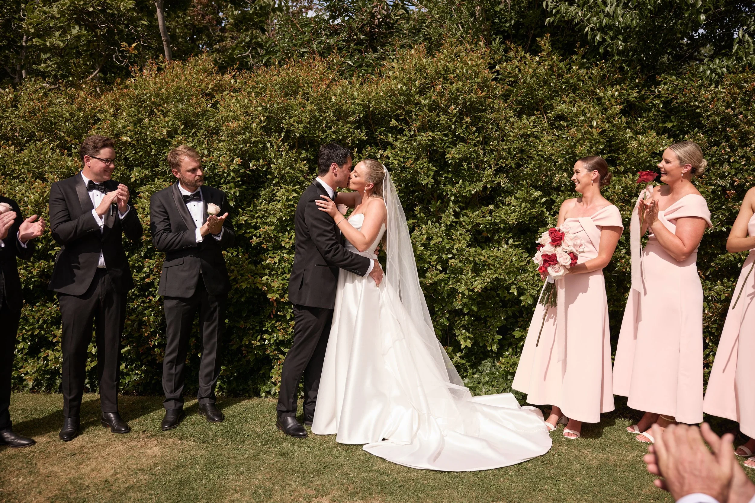 An extravagant wedding ceremony outdoors with the bride and groom kissing, surrounded by bridesmaids and groomsmen clapping.