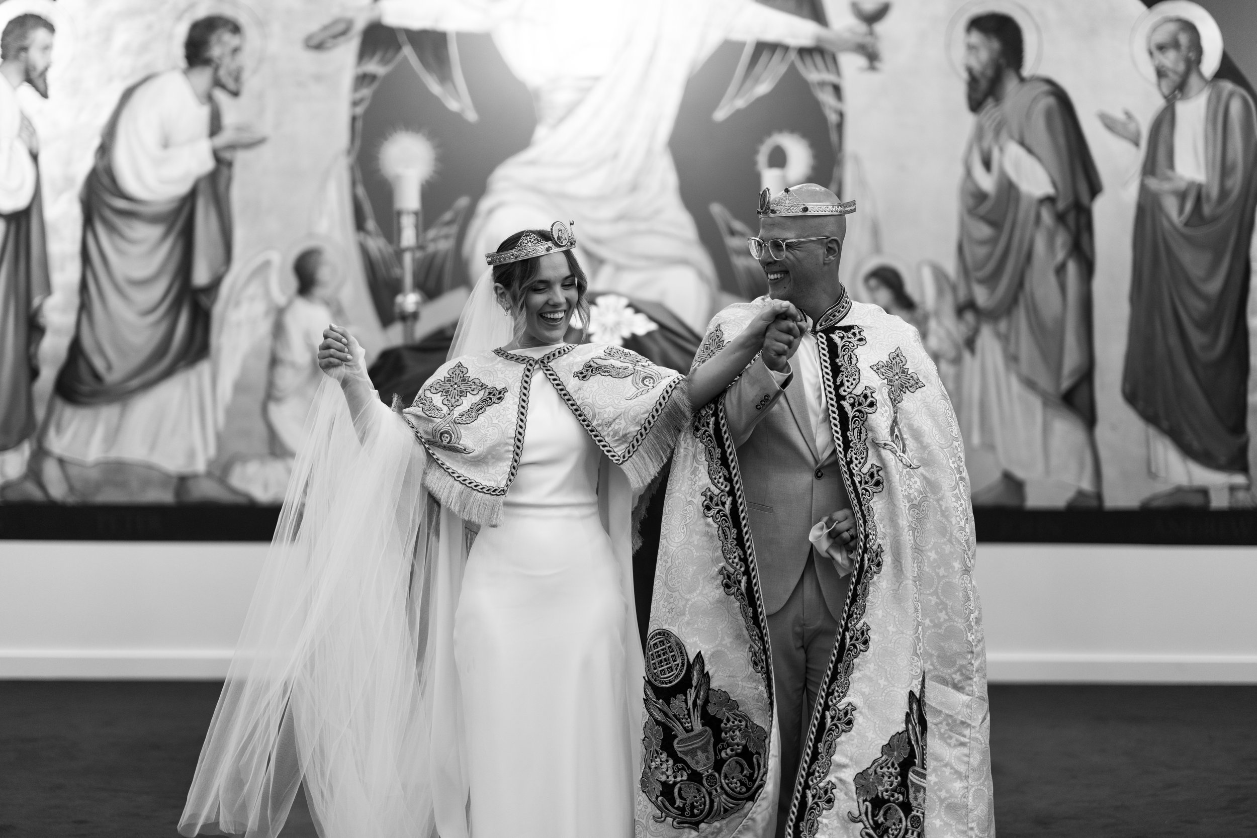 A black and white photo of a couple in traditional wedding attire, possibly participating in an Eastern Orthodox ceremony, wearing crowns and capes, walking hand in hand in front of a religious mural.