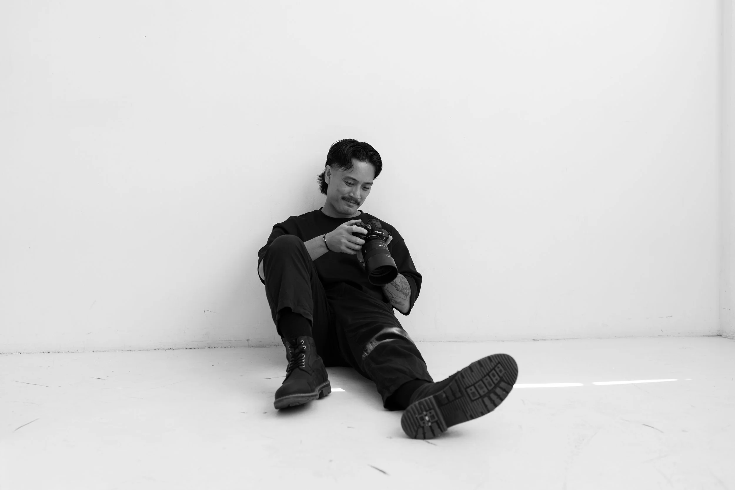 A man with a mustache and dark hair, wearing a black shirt and boots, is sitting on the floor against a plain white wall, looking at a camera he is holding.