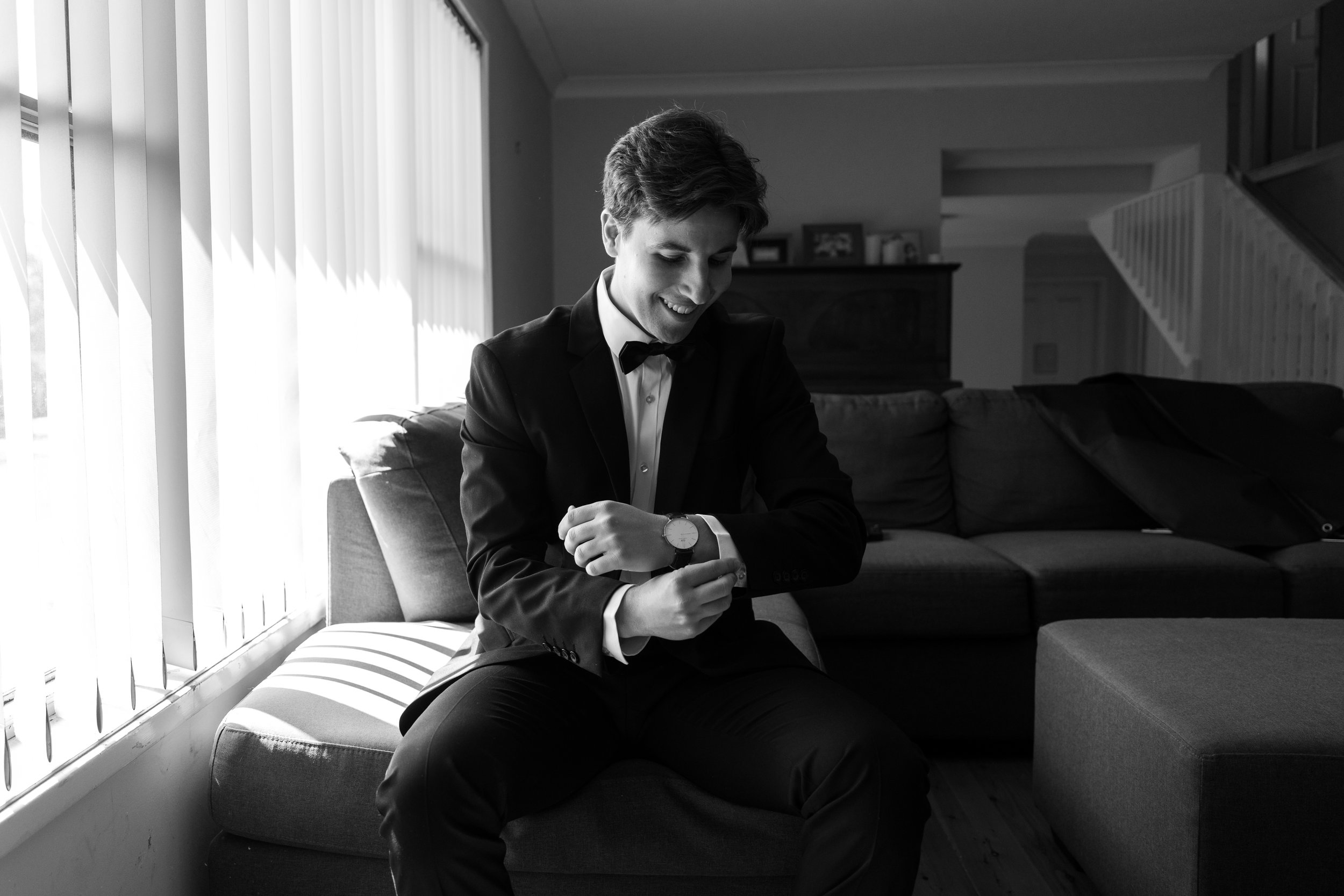 Man in a suit checking his watch while sitting on a couch in a living room, with natural light filtering through blinds.