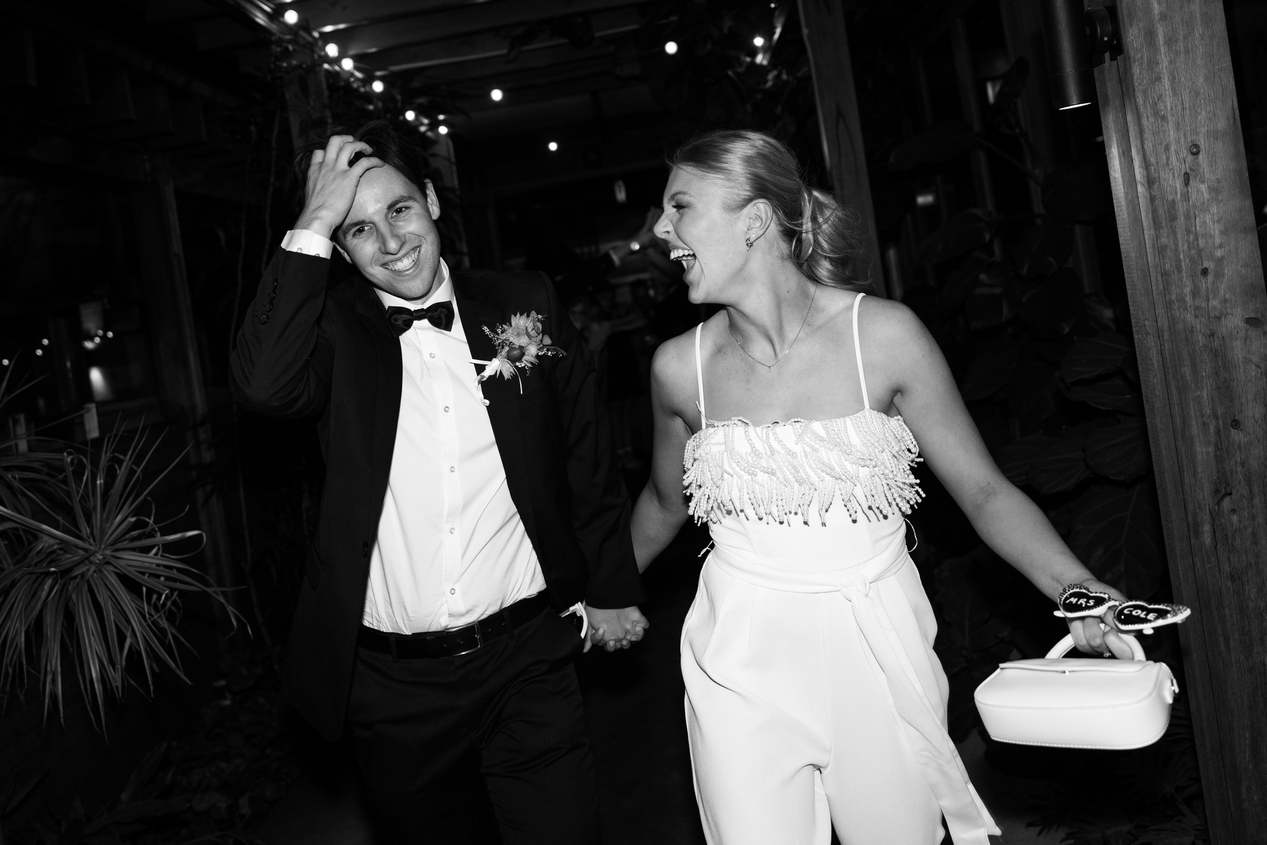 A happy couple walking hand in hand, dressed in formal attire. The man is in a suit with a boutonniere, and the woman is in a stylish jumpsuit, holding a small purse and a pair of glasses. They appear to be at a celebration, smiling and laughing toge
