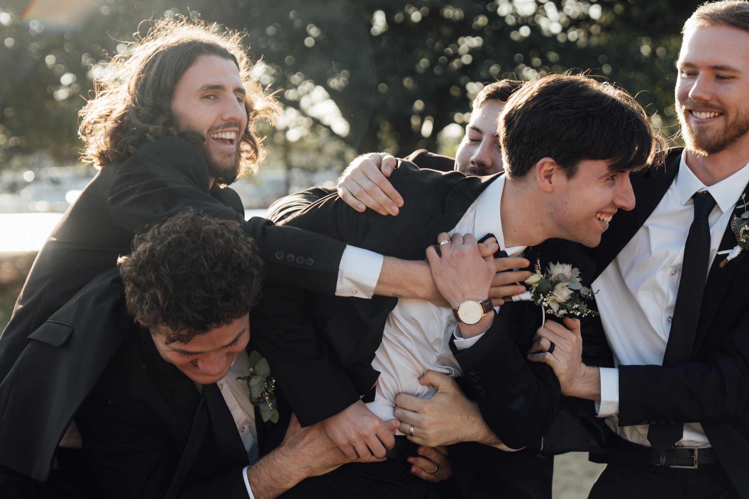 Group of groomsmen in suits playfully interacting outdoors, wearing boutonnieres.