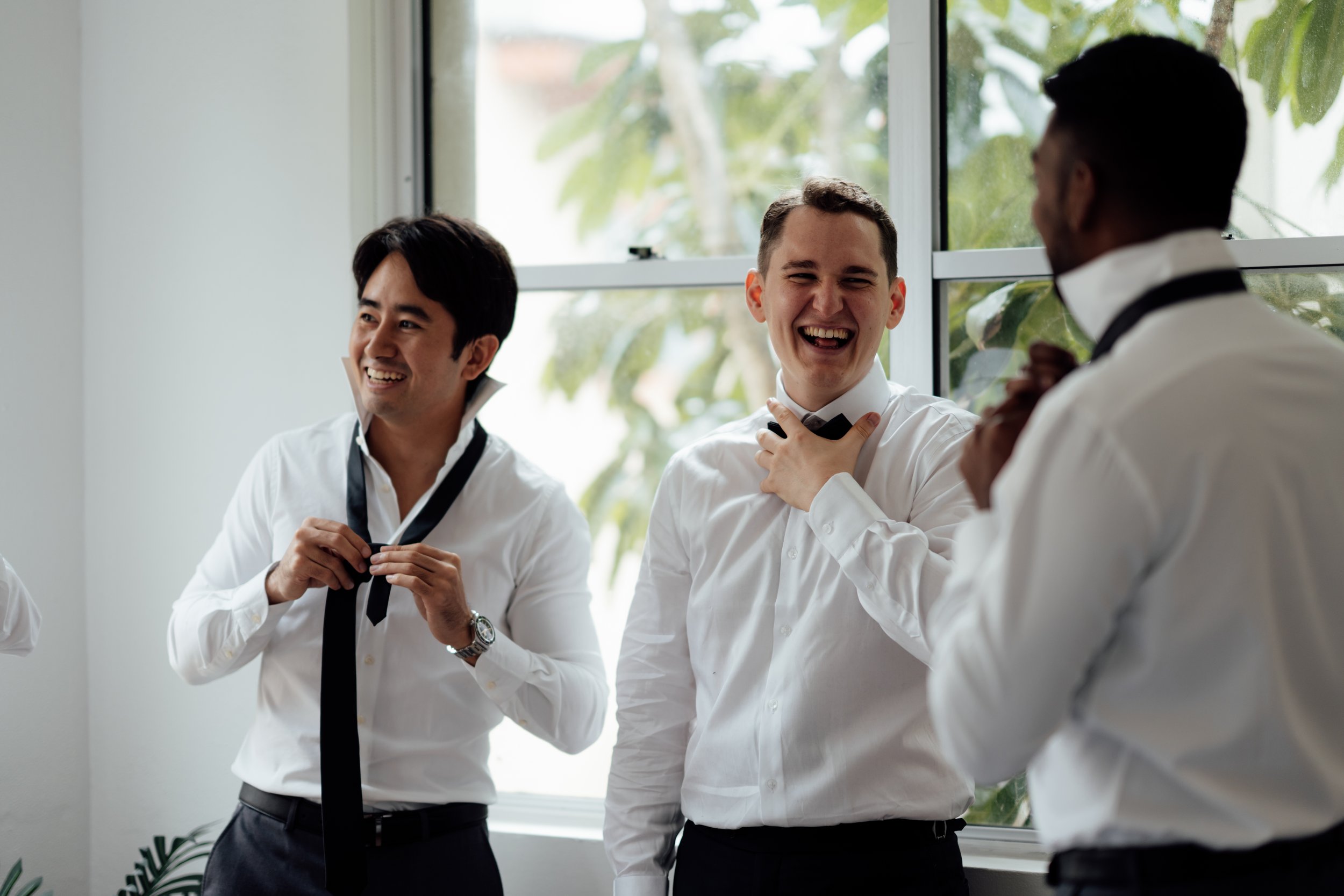 Three men in white shirts, smiling and adjusting black ties, stand near a window.
