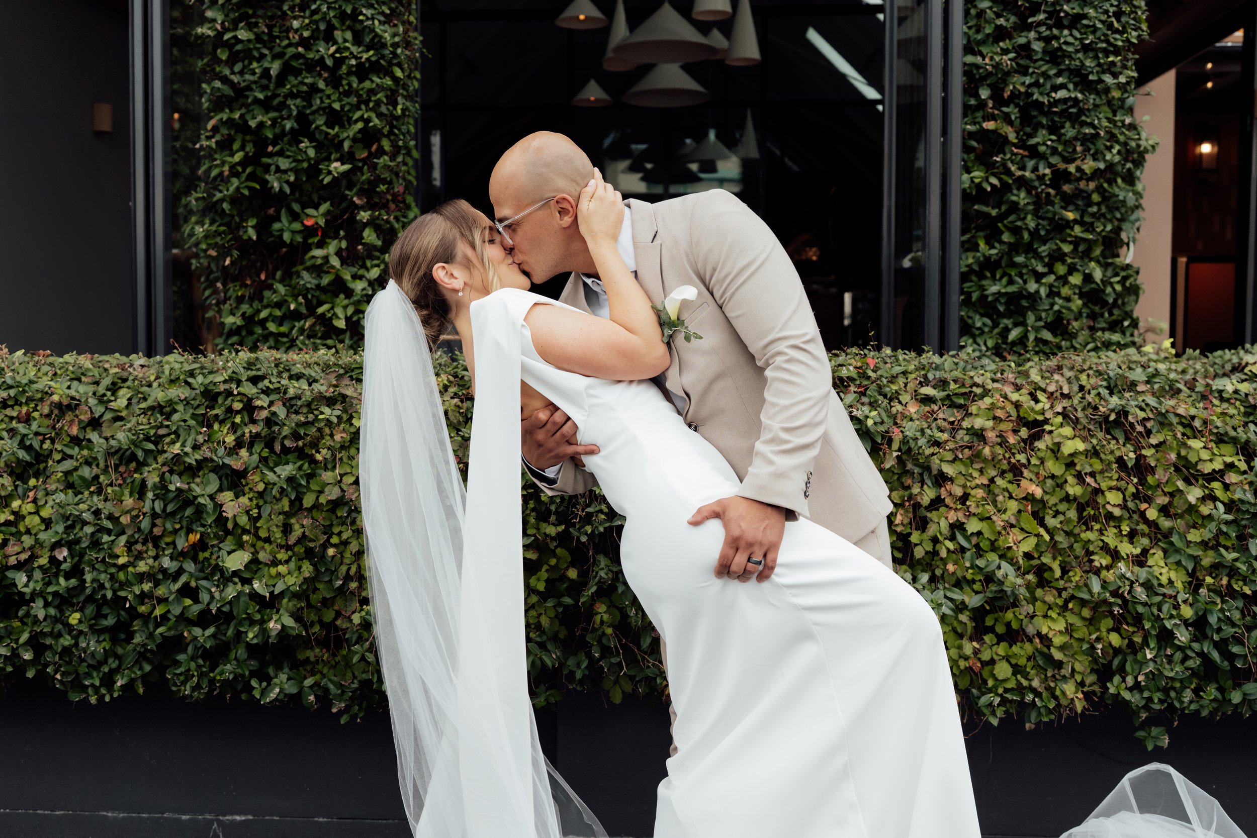 Bride in white wedding dress and groom in beige suit kissing in front of green foliage wall.