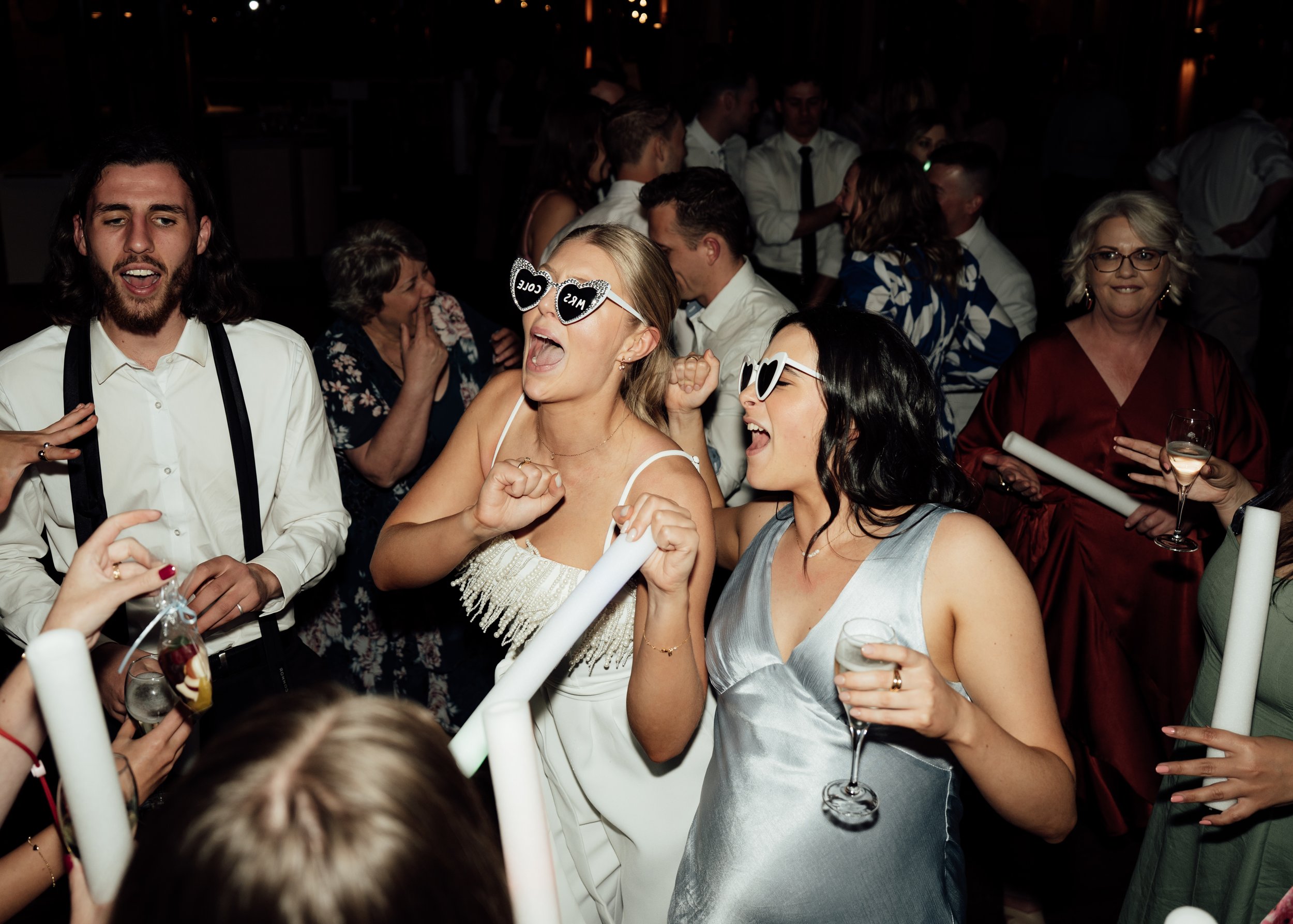People celebrating at a party, wearing sunglasses, holding drinks and glow sticks, and dancing.