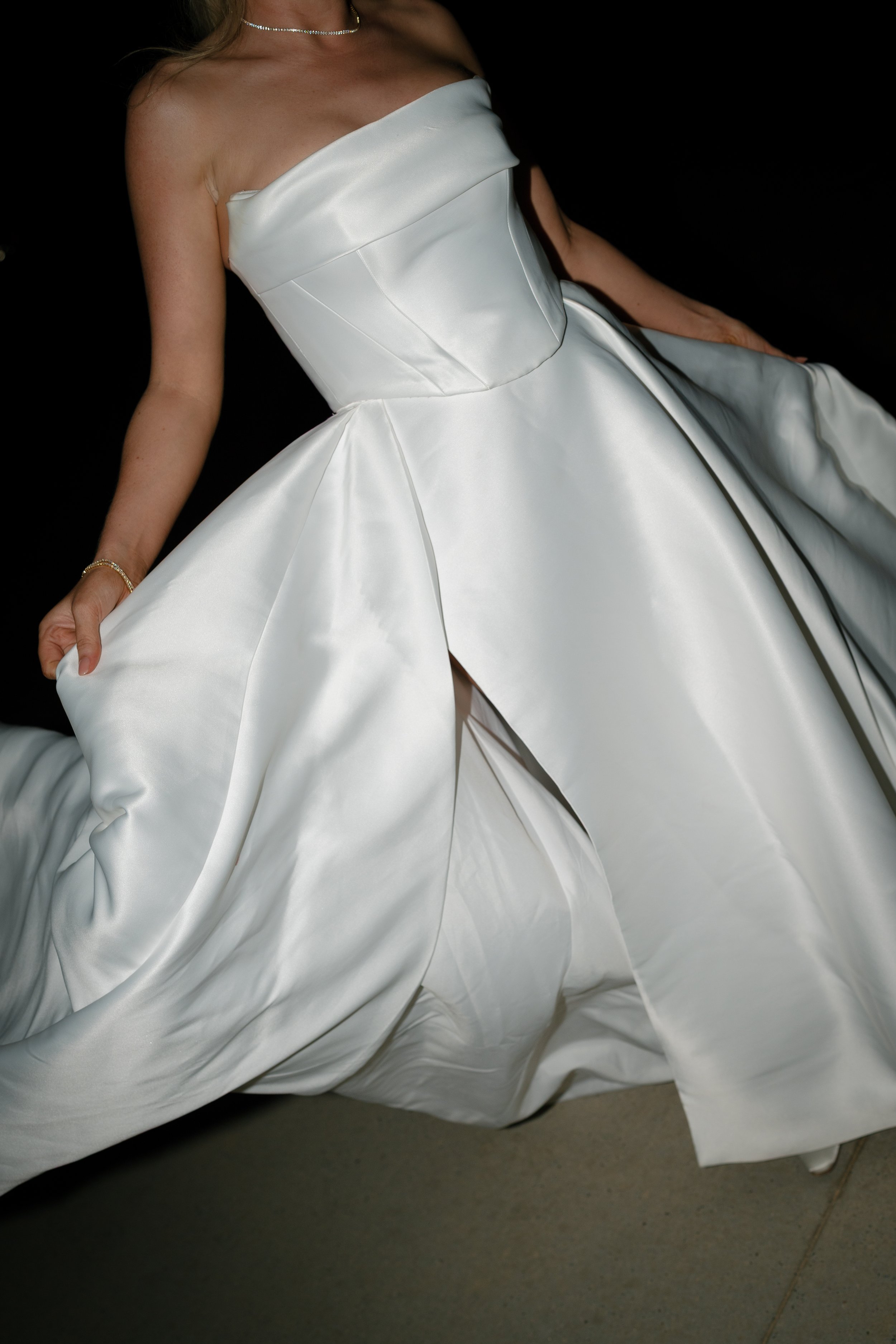 A woman wearing a strapless white satin gown with a fitted bodice and a full skirt. She is accessorized with a pearl necklace, a bracelet, and has long, wavy hair.