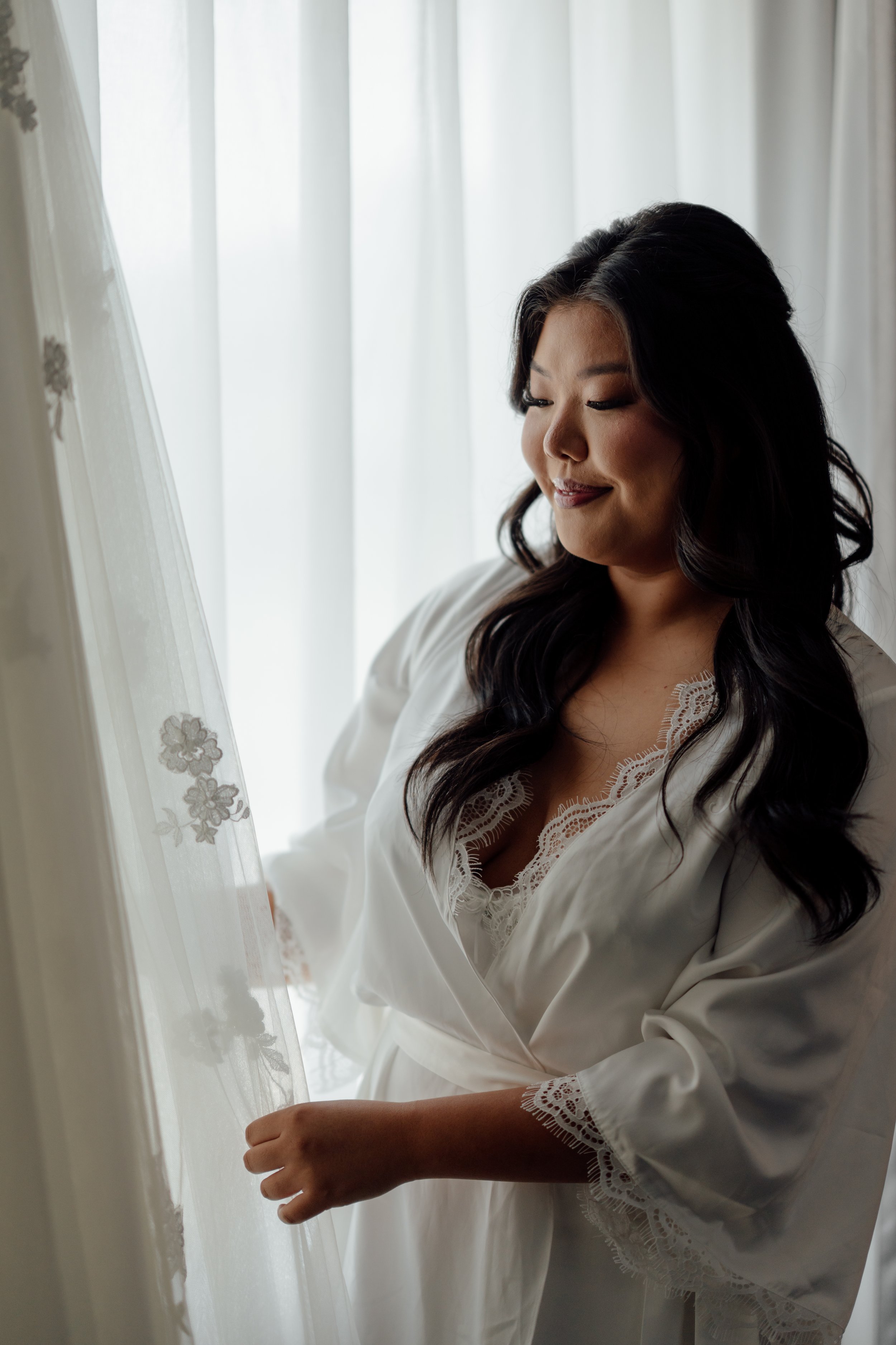 Woman in white lace robe looking at delicate sheer curtains with floral patterns.