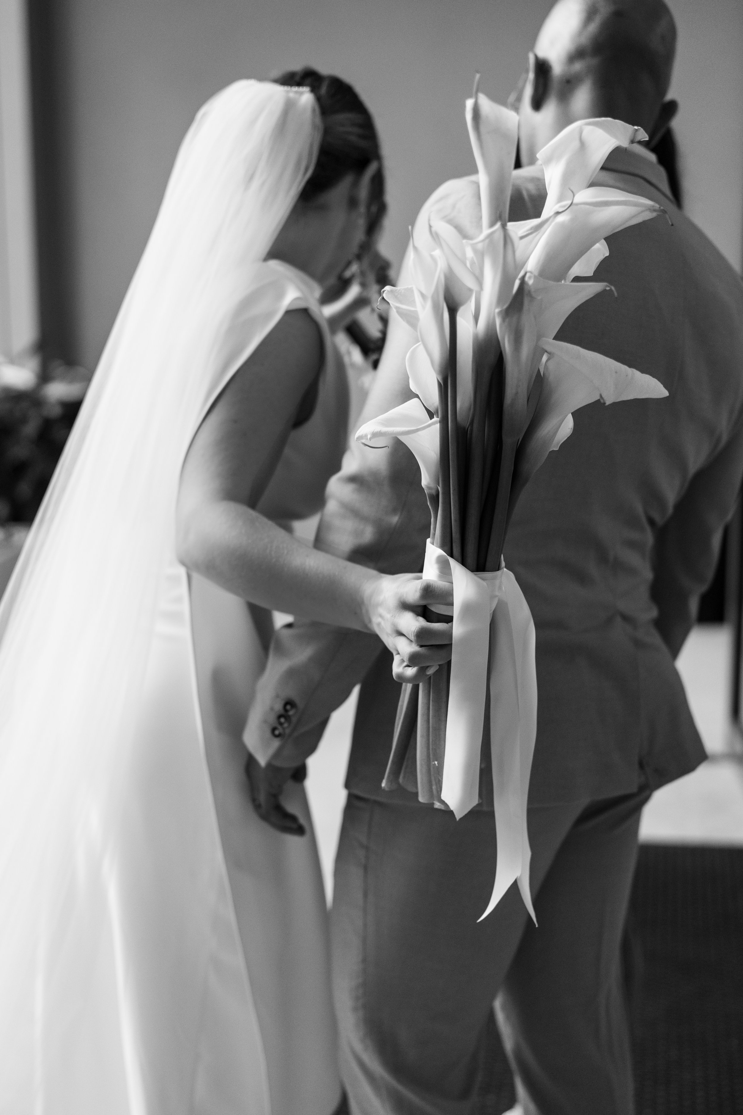A bride in a wedding dress with a veil holding a bouquet of white calla lilies, standing next to a person in a suit, shot in black and white.