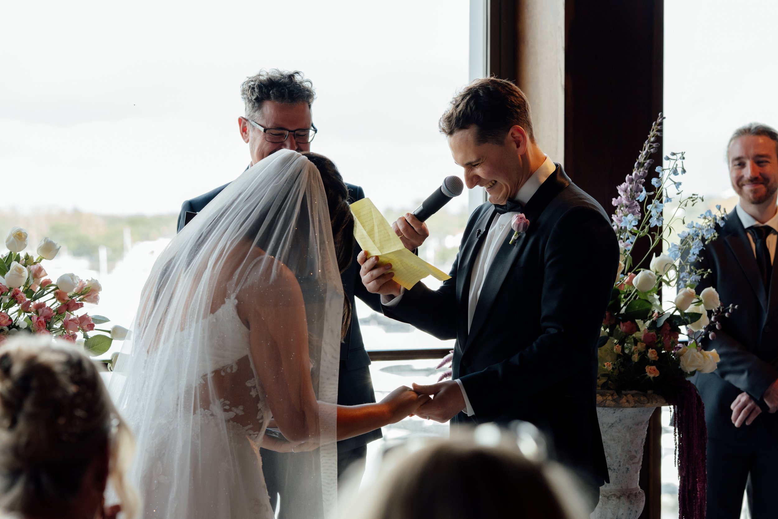 Bride and groom exchanging vows during a wedding ceremony, with a man holding a microphone and witnesses nearby, surrounded by floral arrangements.