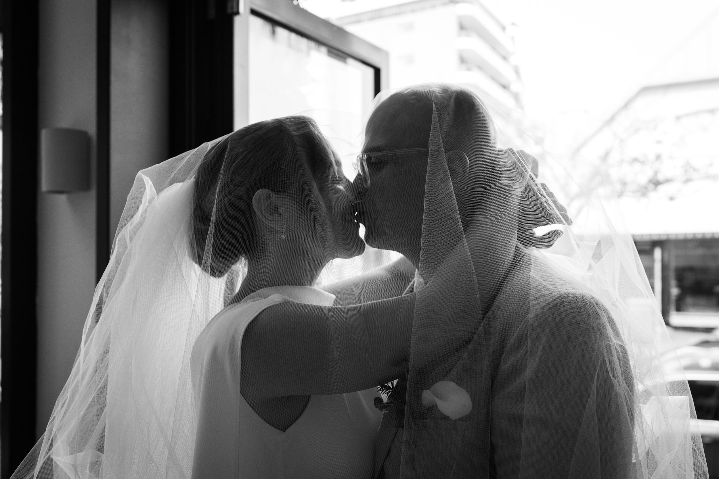 Bride and groom kissing under a veil in black and white photo.