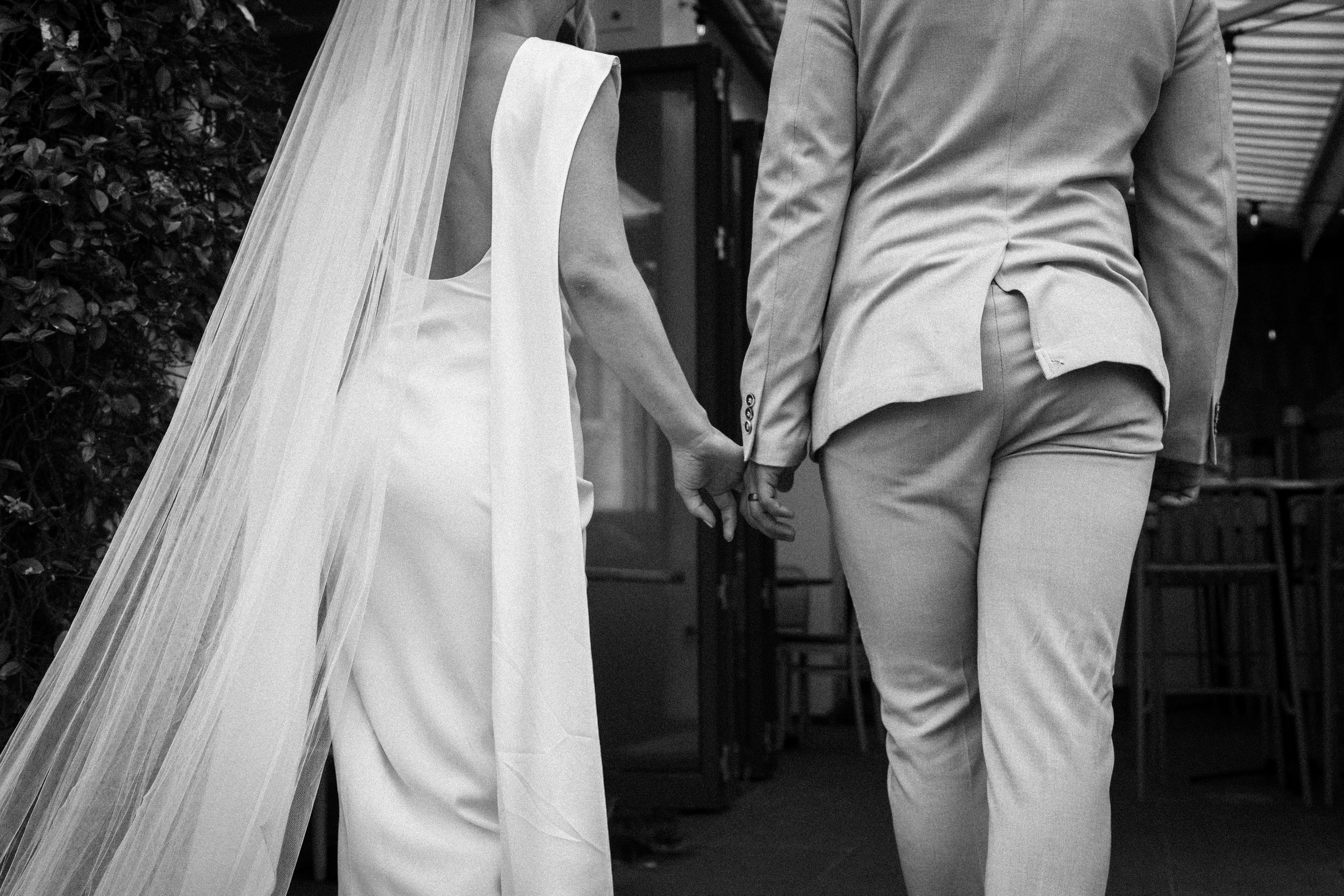 Black and white photo of a couple walking away, holding hands, with a bride in a gown and veil, and a groom in a suit.