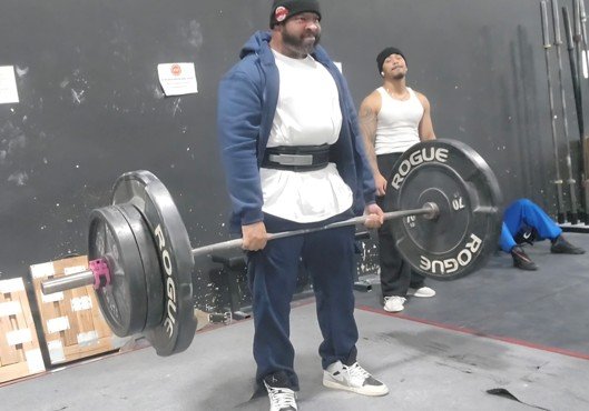 A man wearing a blue hoodie, white shirt, and black weightlifting belt is lifting a barbell with large weights in a gym. A person in a white tank top and black cap stands behind him.