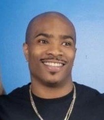 A smiling man with a shaved head, wearing a black shirt and gold chain, against a blue background.