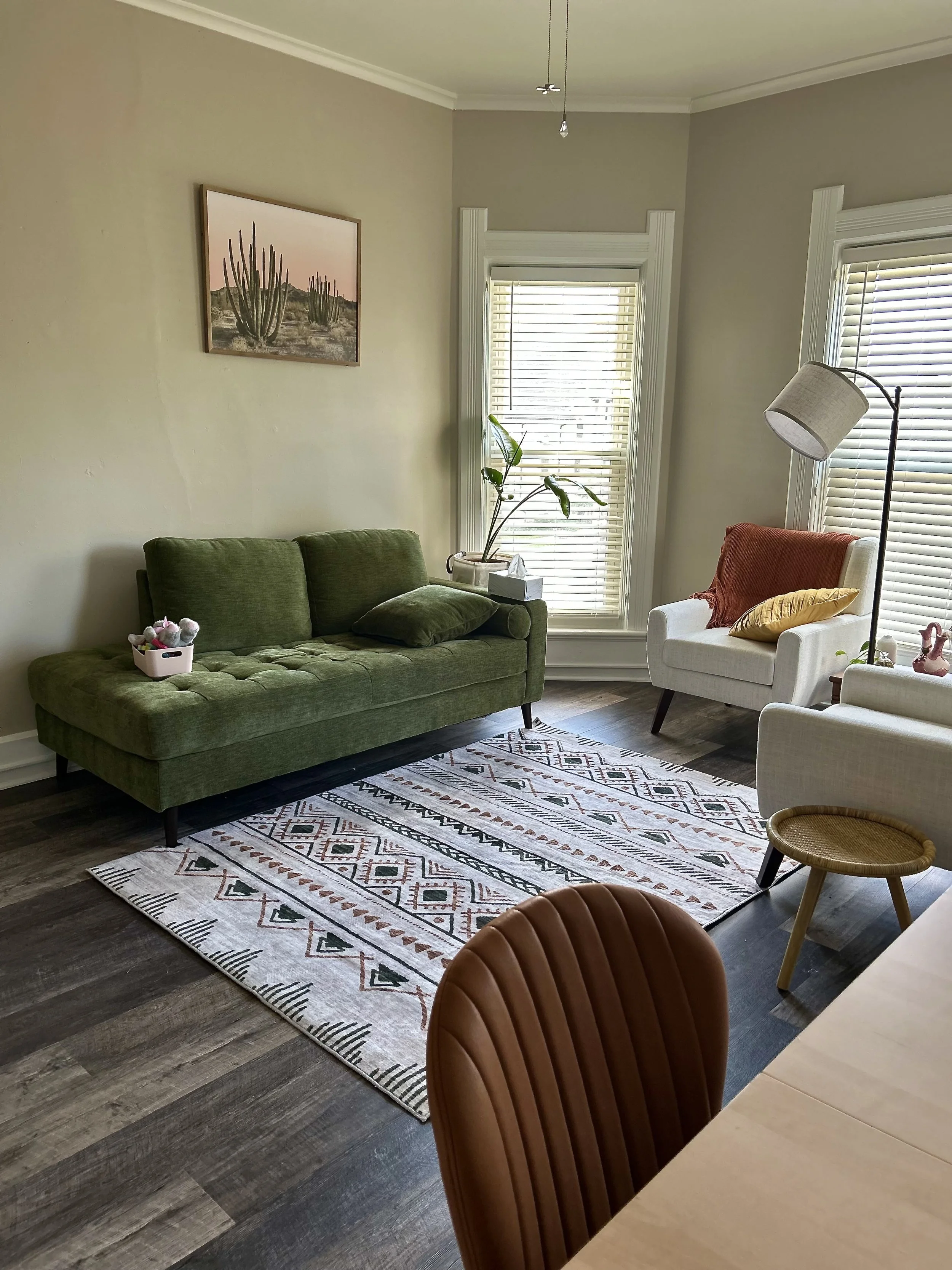 Living room with a green sofa, white armchair, a patterned rug, wooden floor, walls, windows with blinds, a floor lamp, and decorative plants and art.
