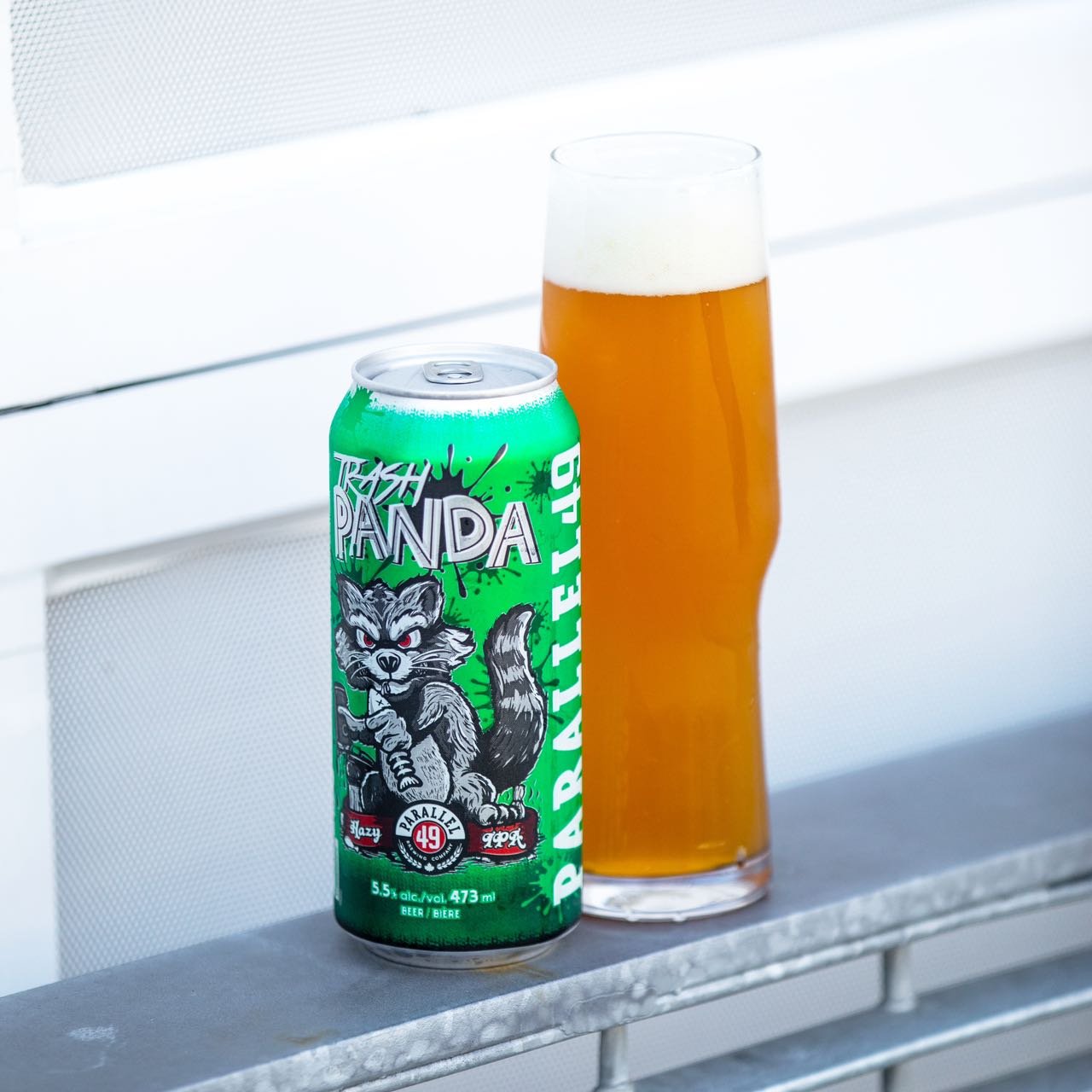 A green can of Parallel 49 Trash Panda beer next to a full tall glass of beer on a metallic surface.
