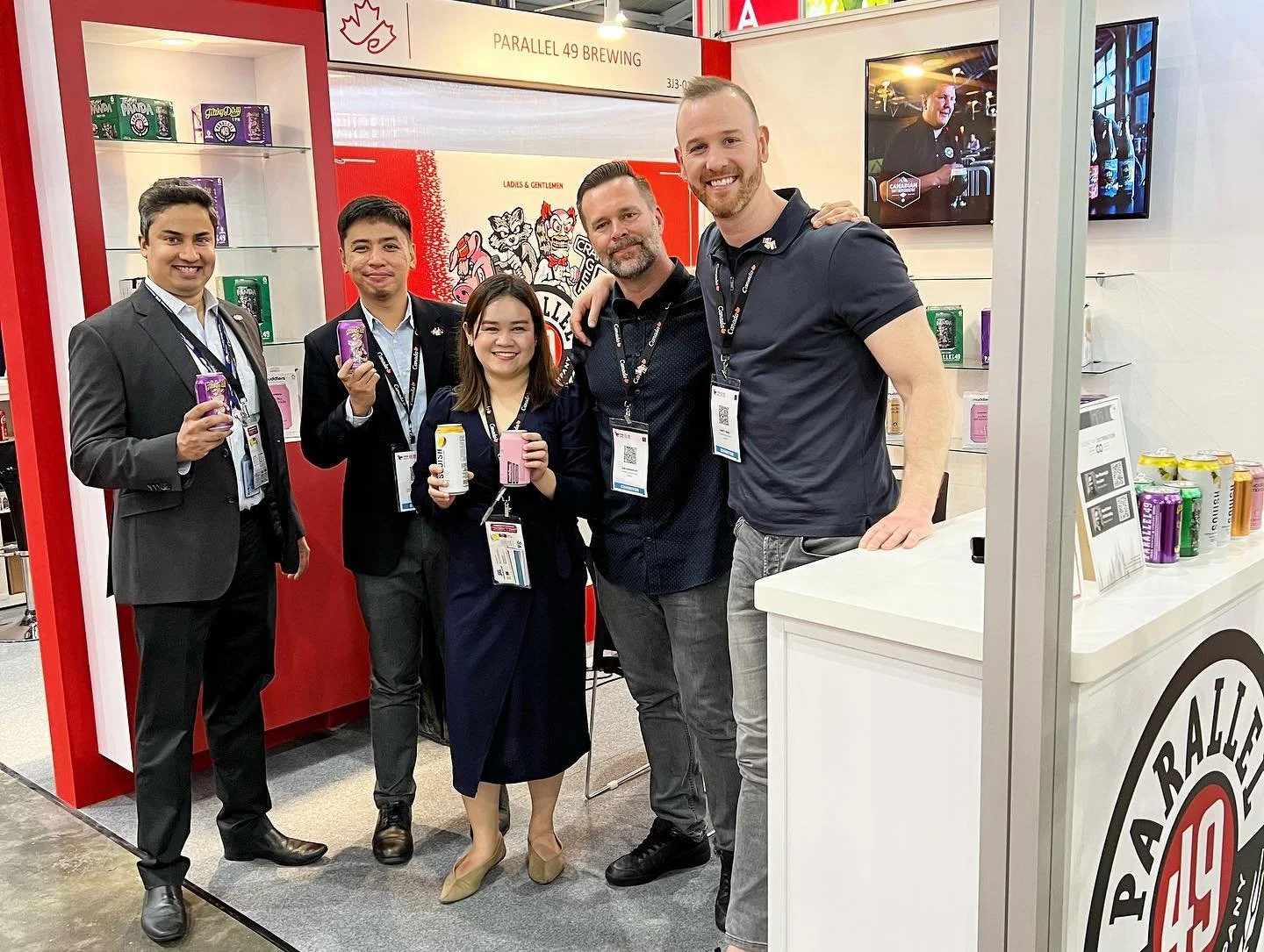 A group of five people standing inside an exhibition booth for Parallel 49 Brewing, holding cans of beverages. The booth features branded signage and a display shelf with various products. They are smiling and wearing name tags, suggesting they are part of a promotional or industry event.