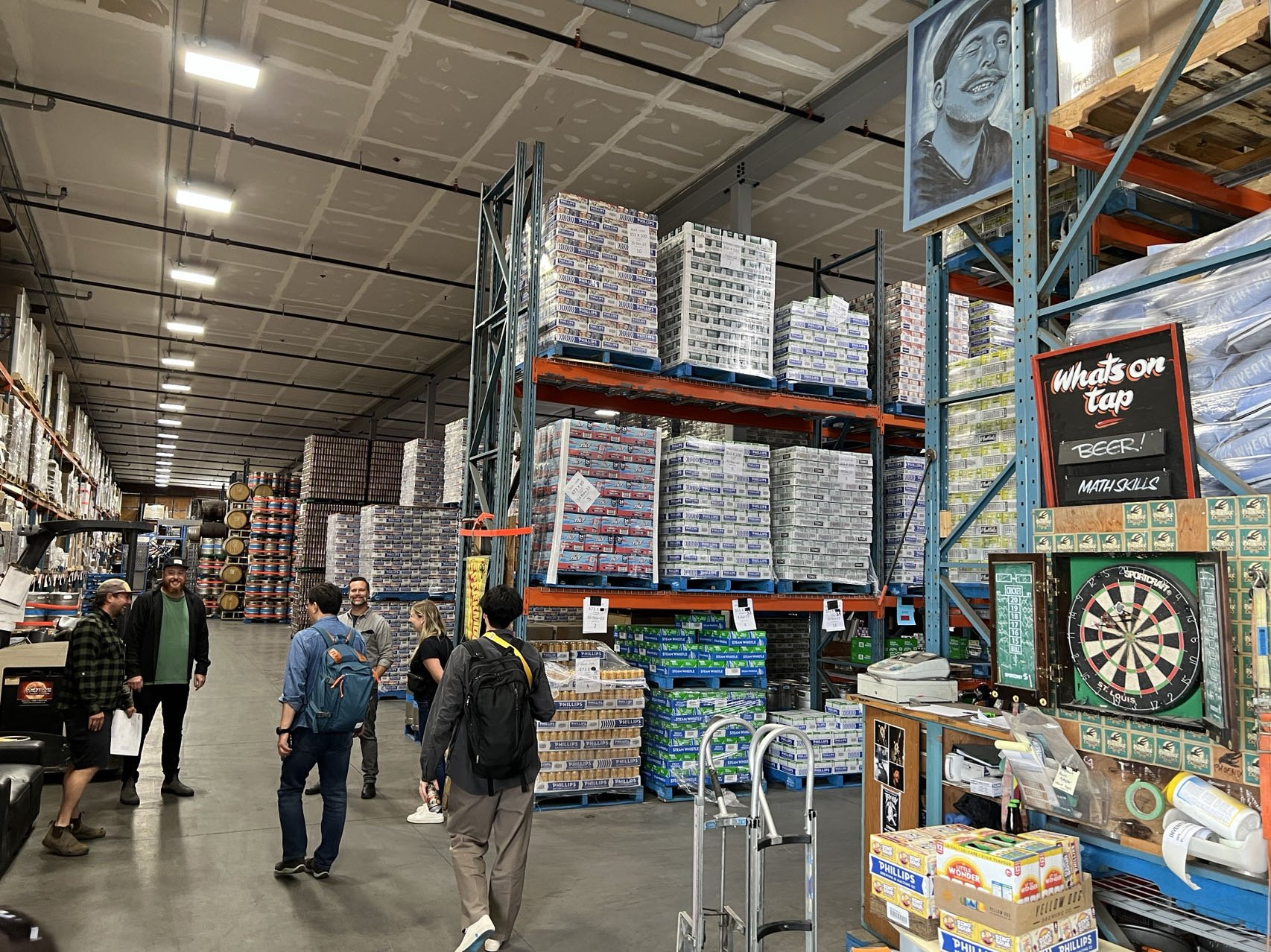 Warehouse interior with stacks of beer boxes on pallets, overhead shelving, a group of people standing, a dartboard on display, and a sign that reads "What's on tap: Beer! Math skills."