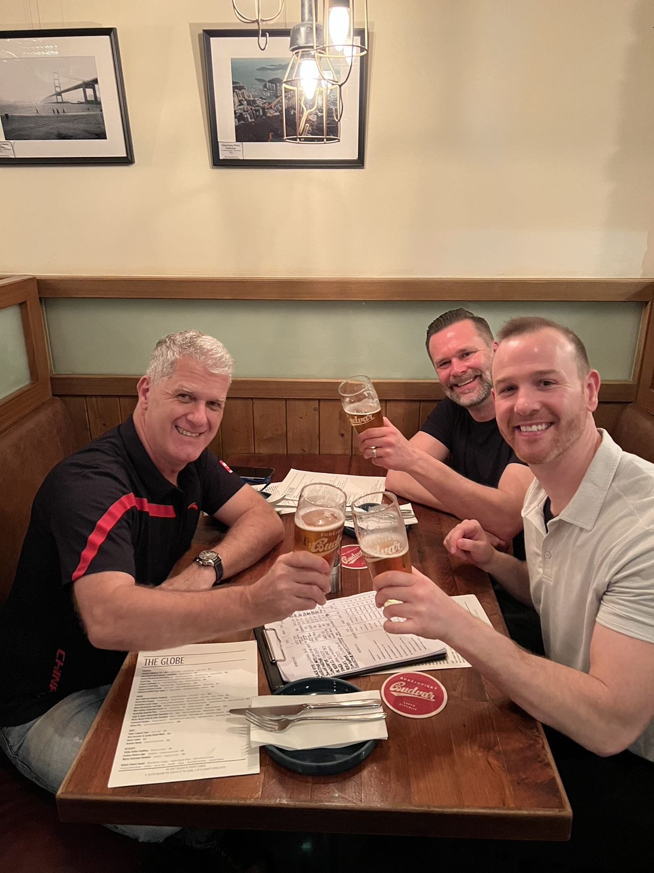 Three men sitting at a restaurant table holding glasses of beer, smiling, with menus on the table, and framed photos on the wall behind them.
