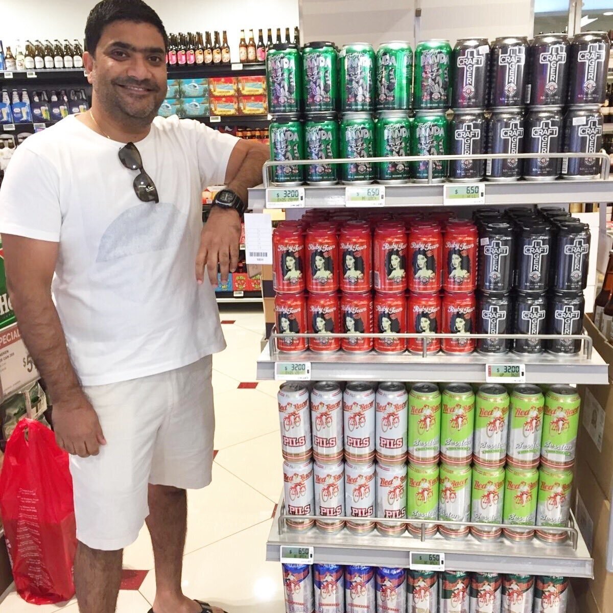 Man standing next to shelves in a store displaying various beer cans, including red, green, black, and white designs. The shelves have price tags, and there are more beverages in the background.