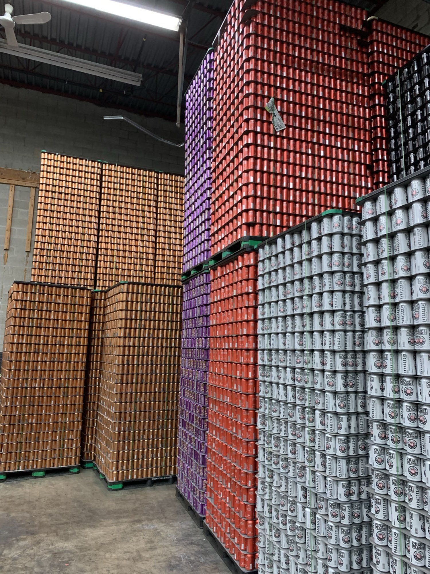Stacks of colorful beverage cans in a warehouse, organized on pallets, featuring varying colors like orange, purple, and white.