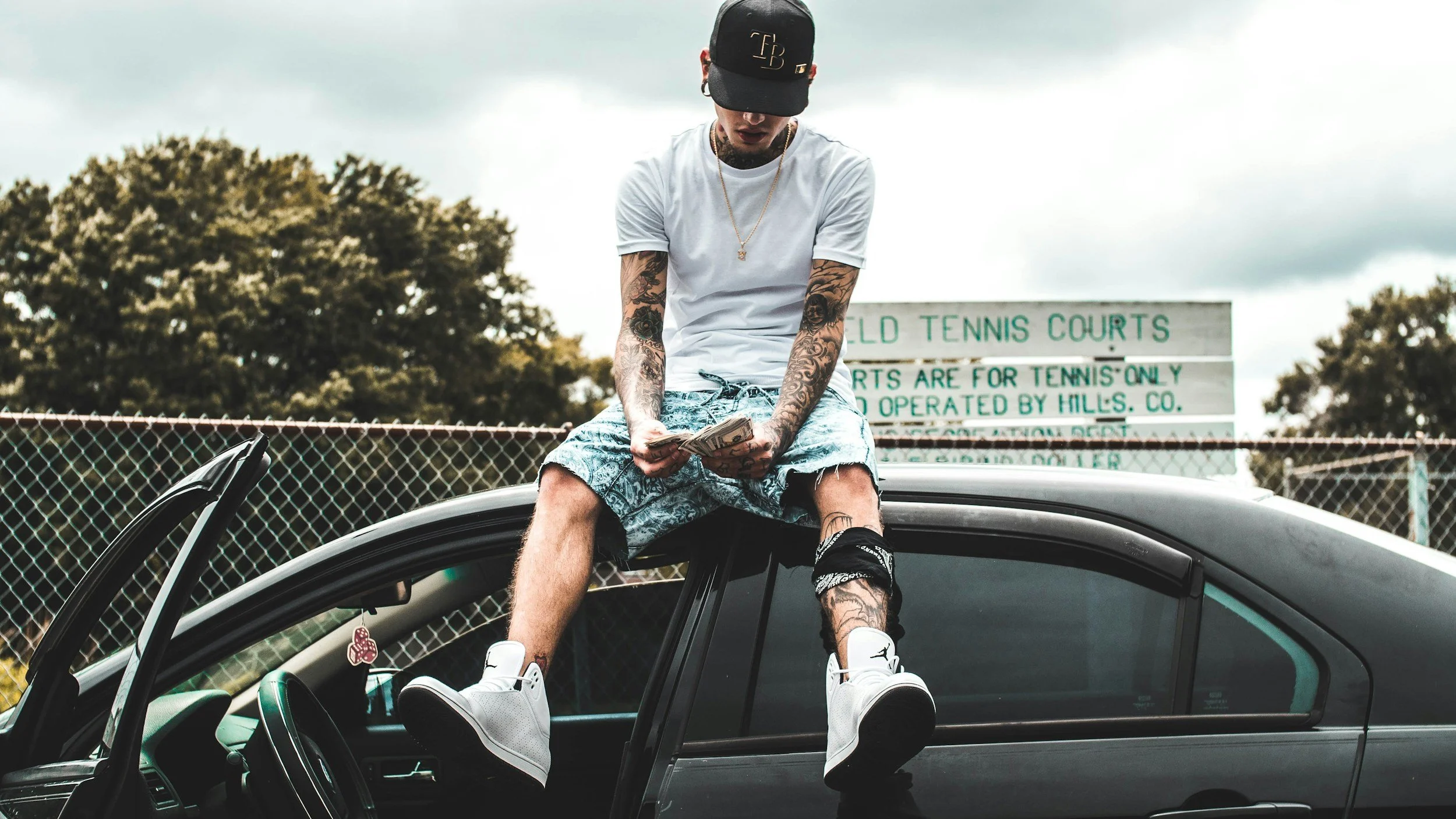 A young man with tattoos wearing a white t-shirt, patterned shorts, and white sneakers, sitting on the open door of a black car while holding cash. Background shows a tennis court fence and a sign that says "Field Tennis Courts".