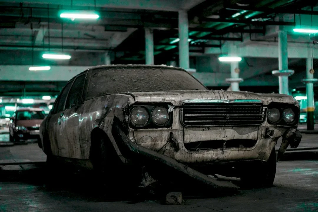 A damaged and dirty classic car with missing parts in a parking garage.