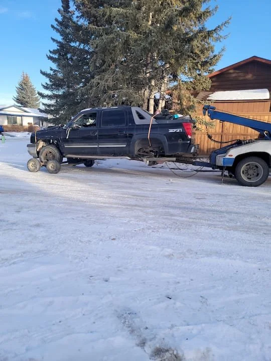 A black pickup truck missing its front wheels is being towed by a flatbed tow truck. The scene is outdoors on a snow-covered street with a house, trees, and a wooden fence in the background.