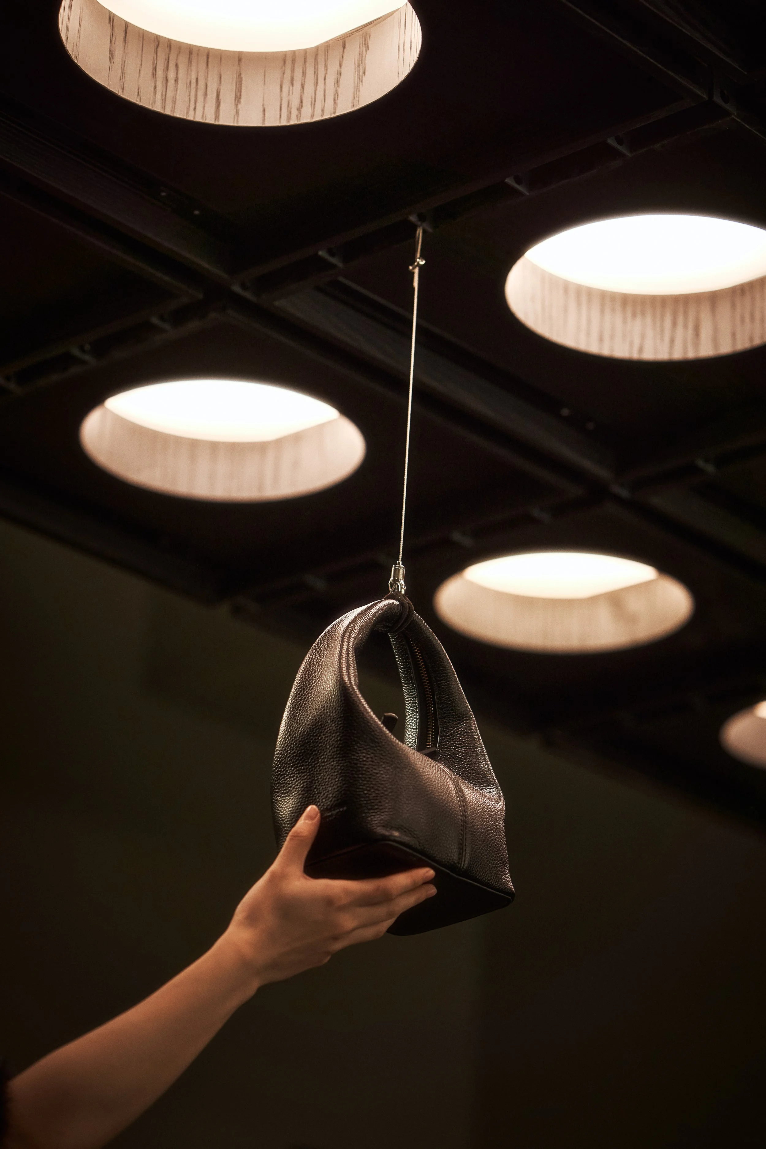 A hand is holding a black handbag hanging from a thin wire attached to the ceiling in a modern interior with circular ceiling lights.