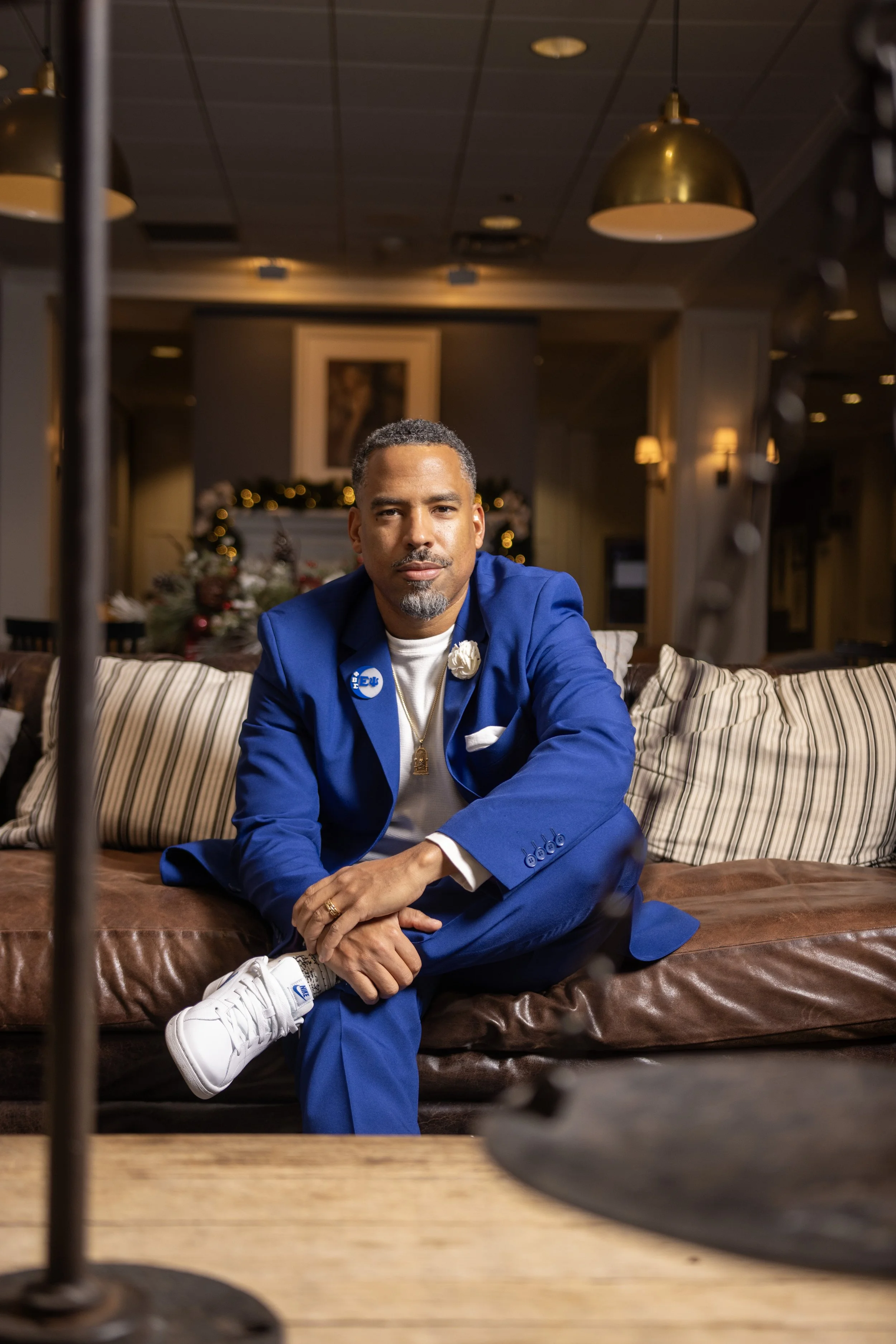 A man in a blue suit sitting on a brown leather couch in a decorated room with holiday ornaments, gold pendant lights, and framed artwork in the background.