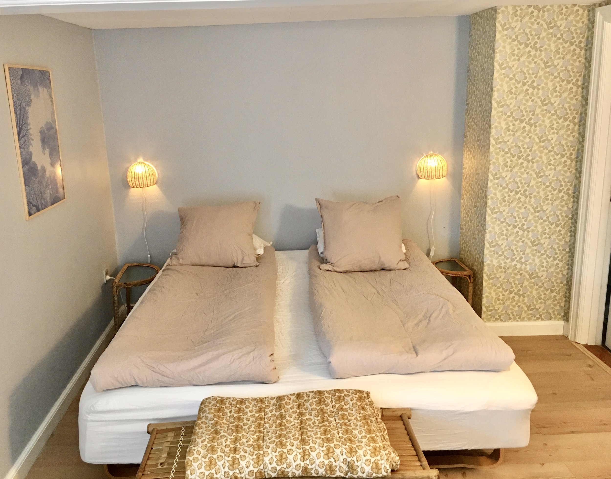 Bedroom with two single beds pushed together, beige bedding, wall-mounted lamps, light gray walls, a patterned accent wall, and a wooden bench at the foot.