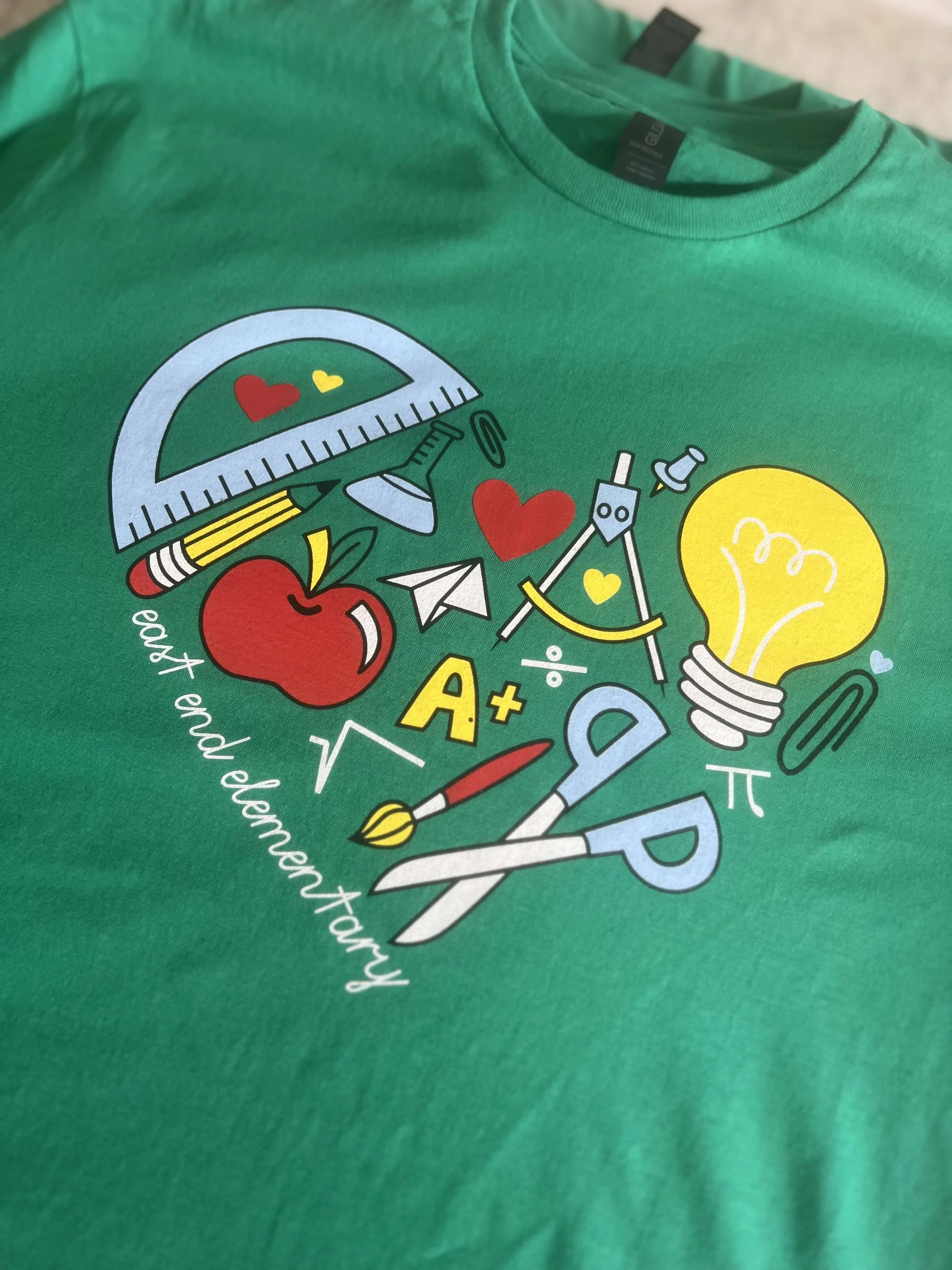Green T-shirt with school-themed graphics, including a protractor, apple, light bulb, scissors, and paintbrush, with text "east end elementary."