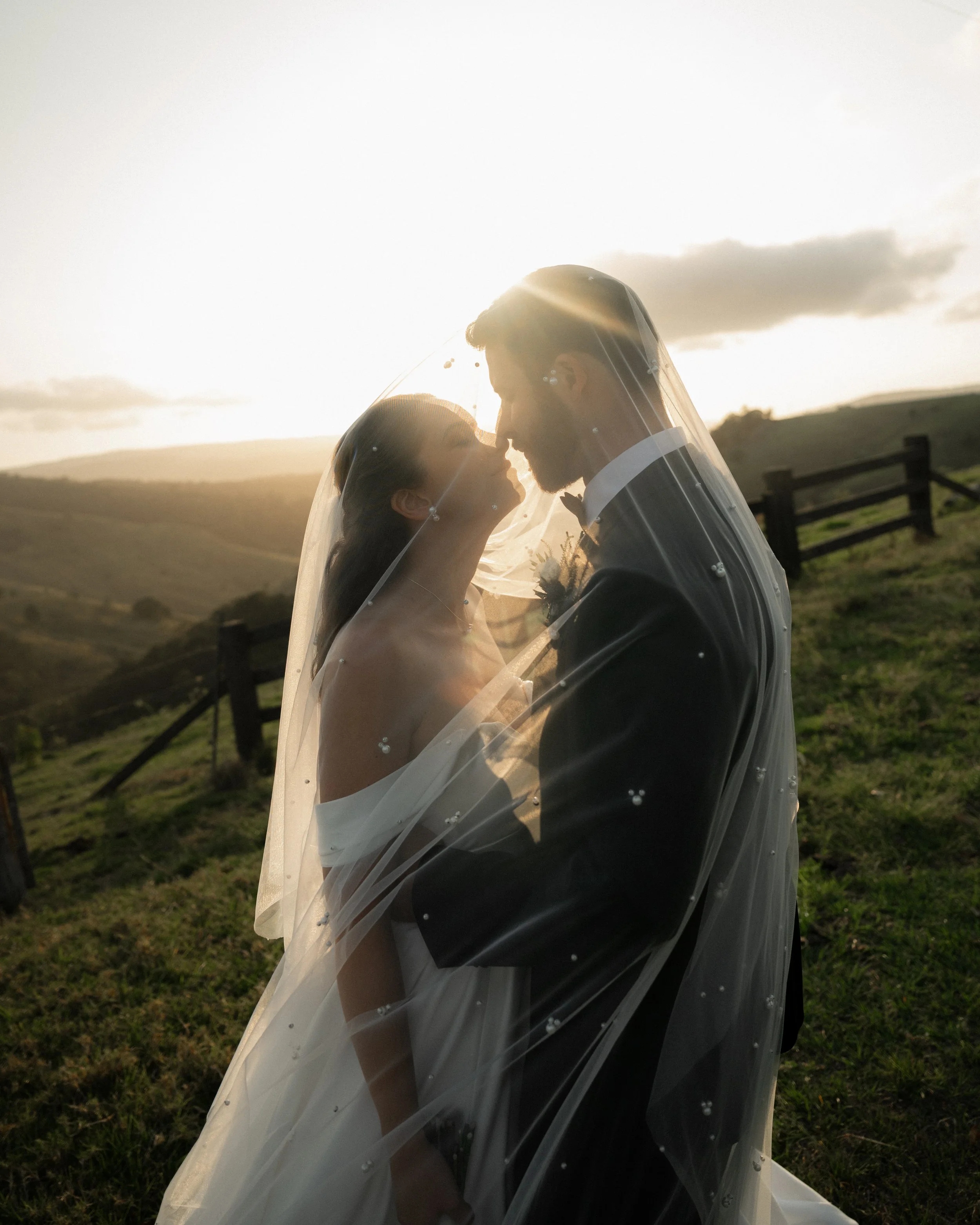 A bride and groom standing close together outdoors at sunset, with the bridal veil covering both of them, touching noses and gazing at each other, in a scenic grassy landscape with hills and a fence.