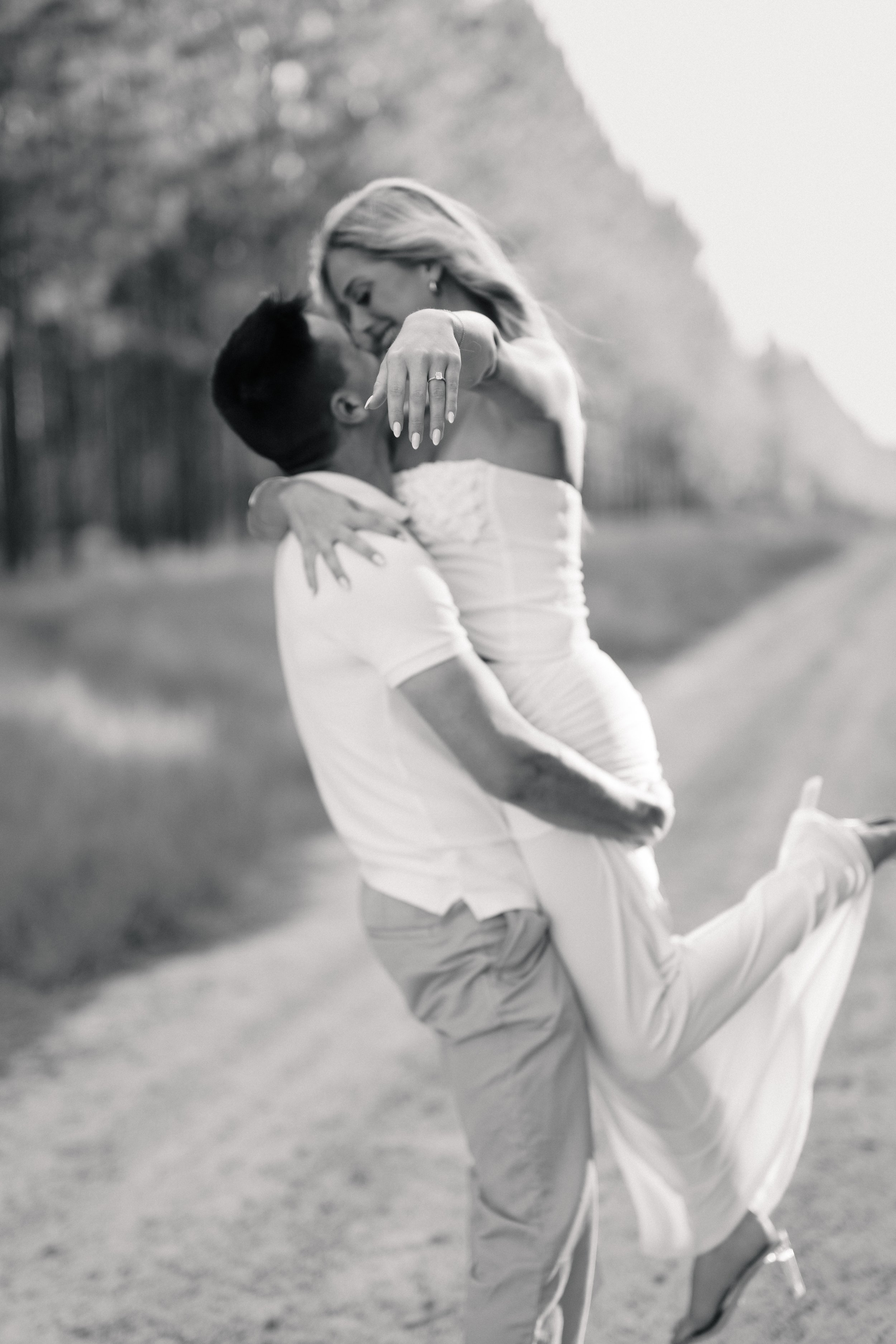 An engaged couple sharing a romantic moment outdoors, with the man lifting the woman in his arms. The woman is showing her engagement ring.