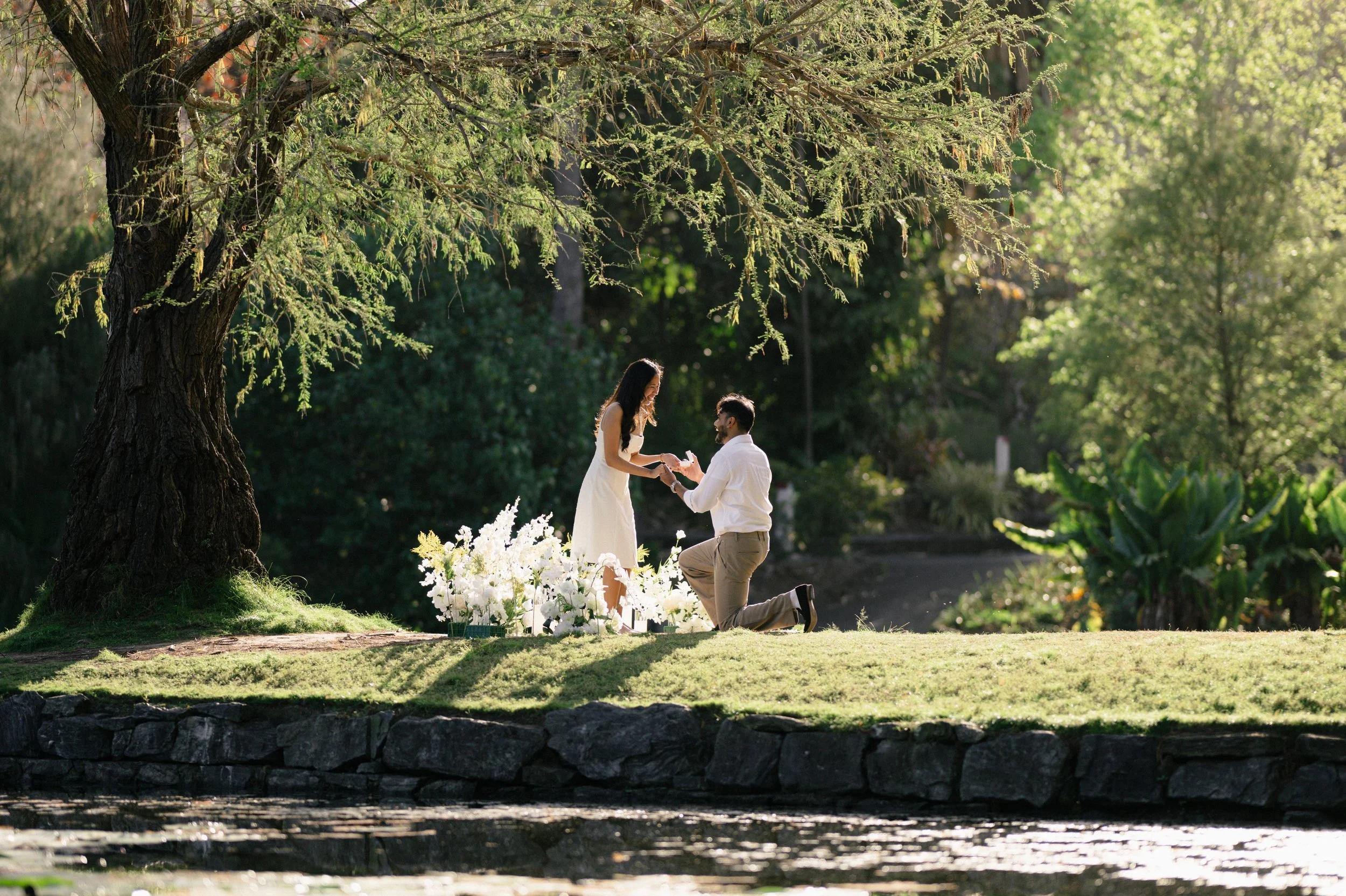 A romantic proposal scene in a park with a man kneeling on one knee offering an engagement ring to a woman standing in front of him. There are white flowers around them, a large tree, and lush greenery in the background.