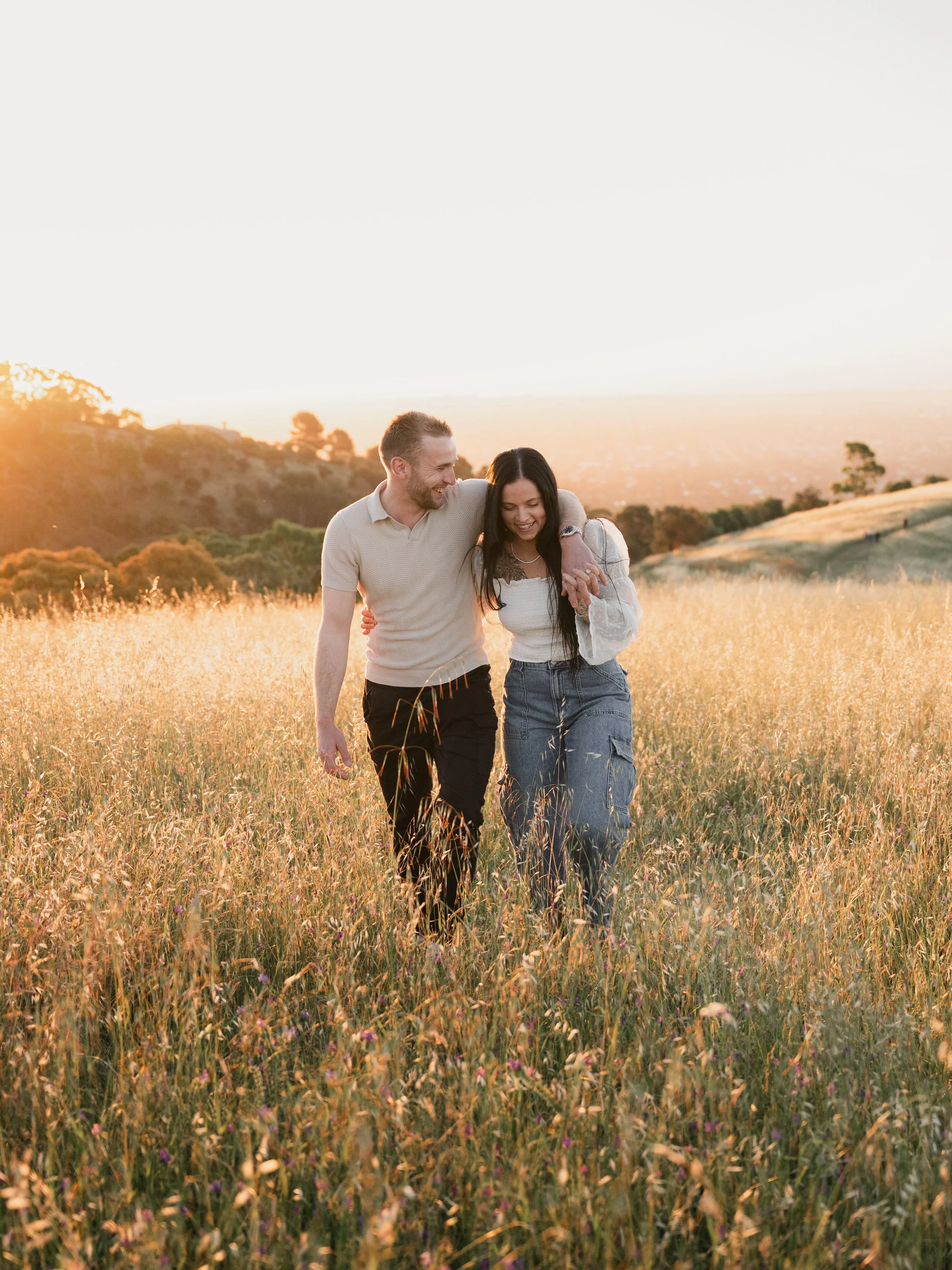 A couple walking arm-in-arm through a grassy field during sunset, smiling and enjoying each other's company.