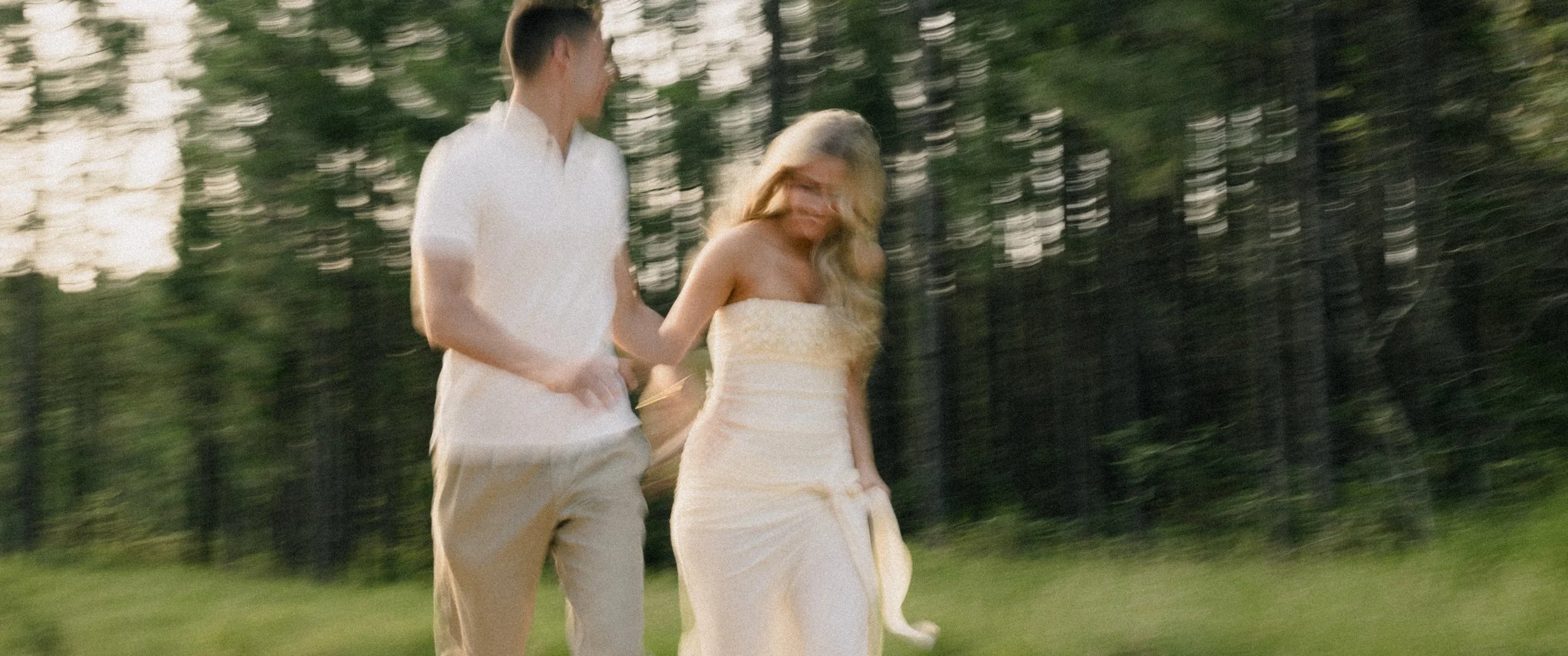 An engaged couple holding hands outdoors in a grassy area with trees in the background, walking together and smiling.