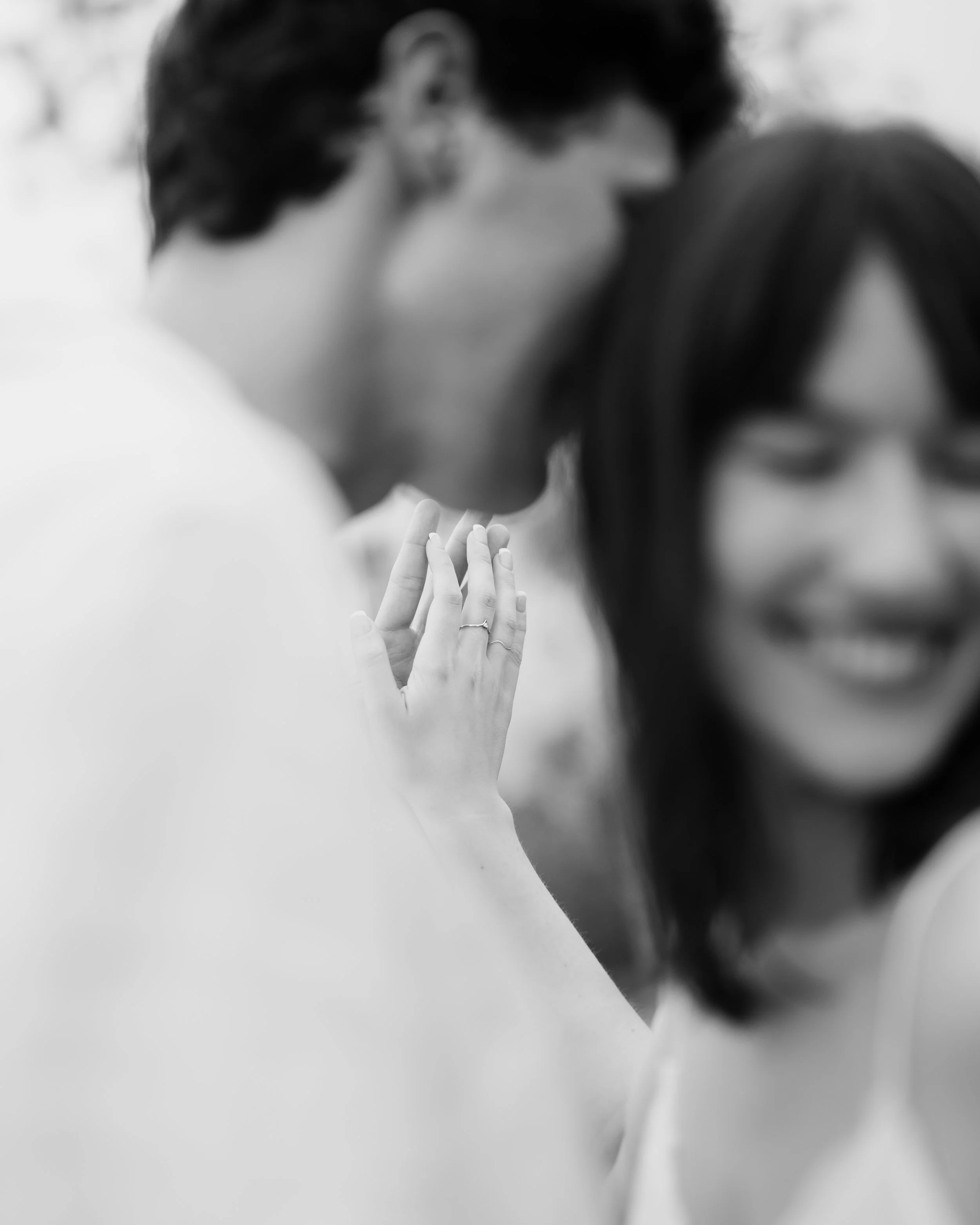 Close-up of a woman's hands with rings, raised and slightly clasped, with a smiling woman and a man behind her, blurred in the background.