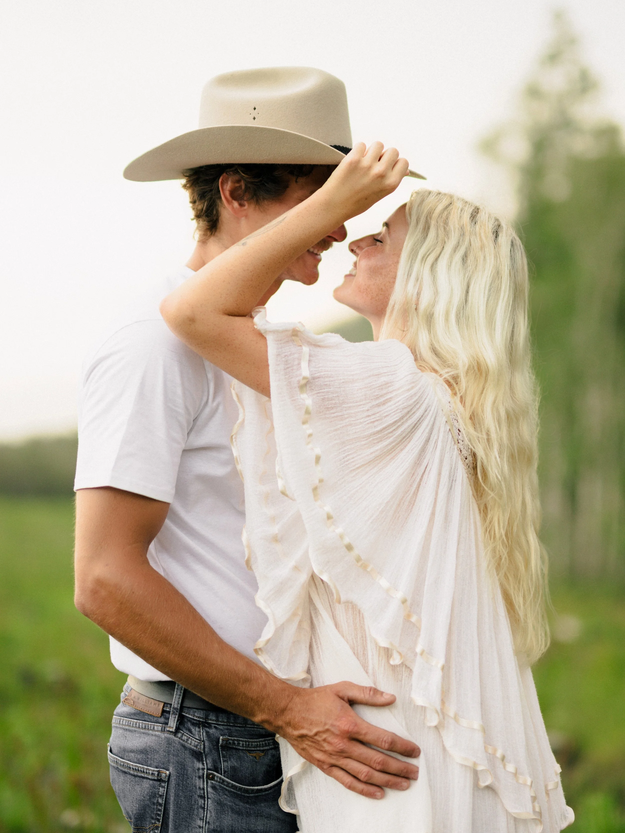 A romantic couple standing close outdoors, smiling and touching foreheads. The man, wearing a beige cowboy hat, a white t-shirt, and jeans, has his arm around the woman. The woman has long, wavy blonde hair and is wearing a light, flowy white dress with ruffled sleeves.