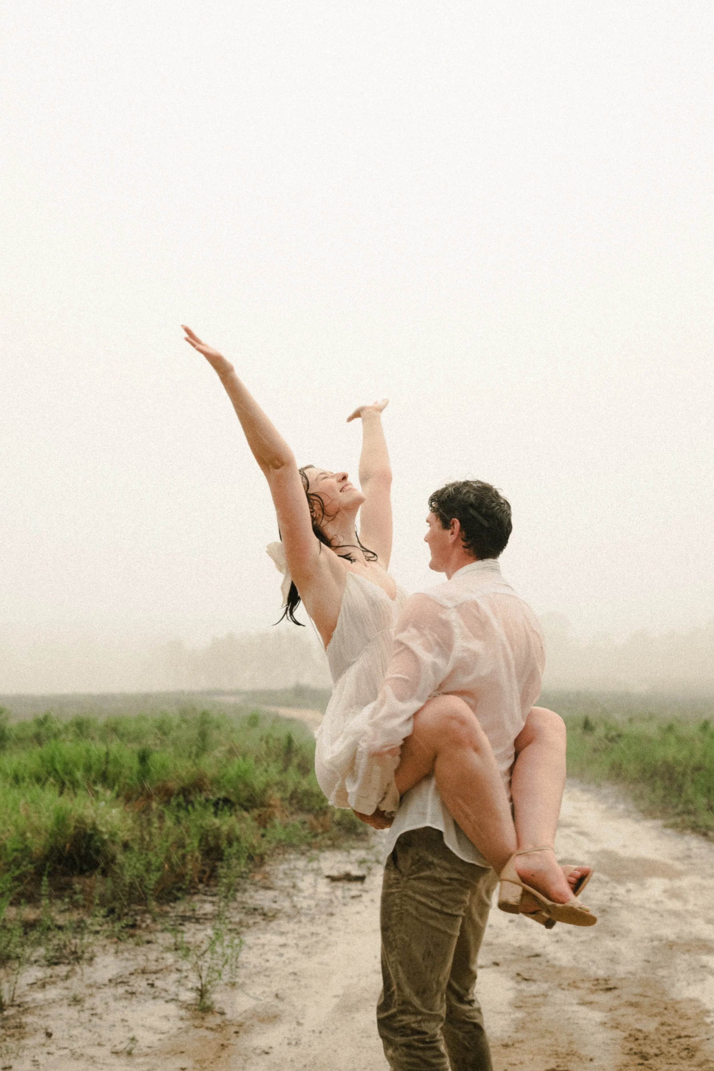 A man is holding a woman in his arms on a dirt path in a grassy outdoor area. The woman has her arms raised and is smiling, wearing a light-colored dress. The man is wearing a light shirt and dark pants, and they both appear happy, enjoying a joyful moment together.