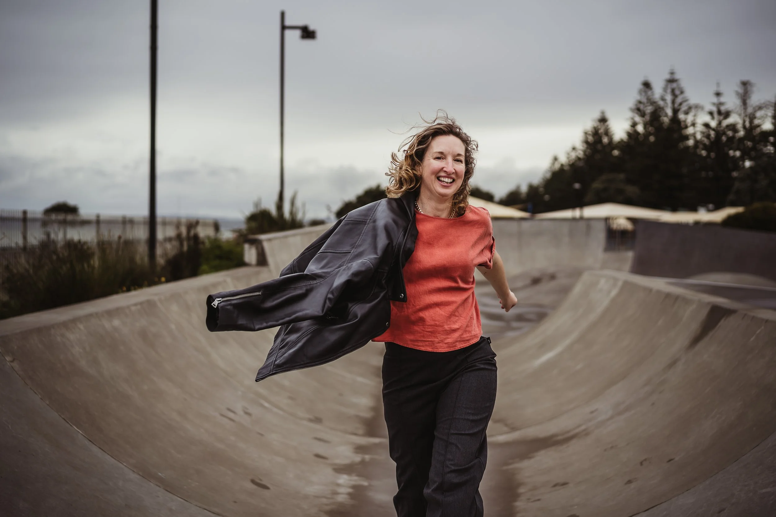 A woman in a red shirt and black pants running through a skate park bowl with a black jacket thrown over her shoulder, smiling under cloudy skies.