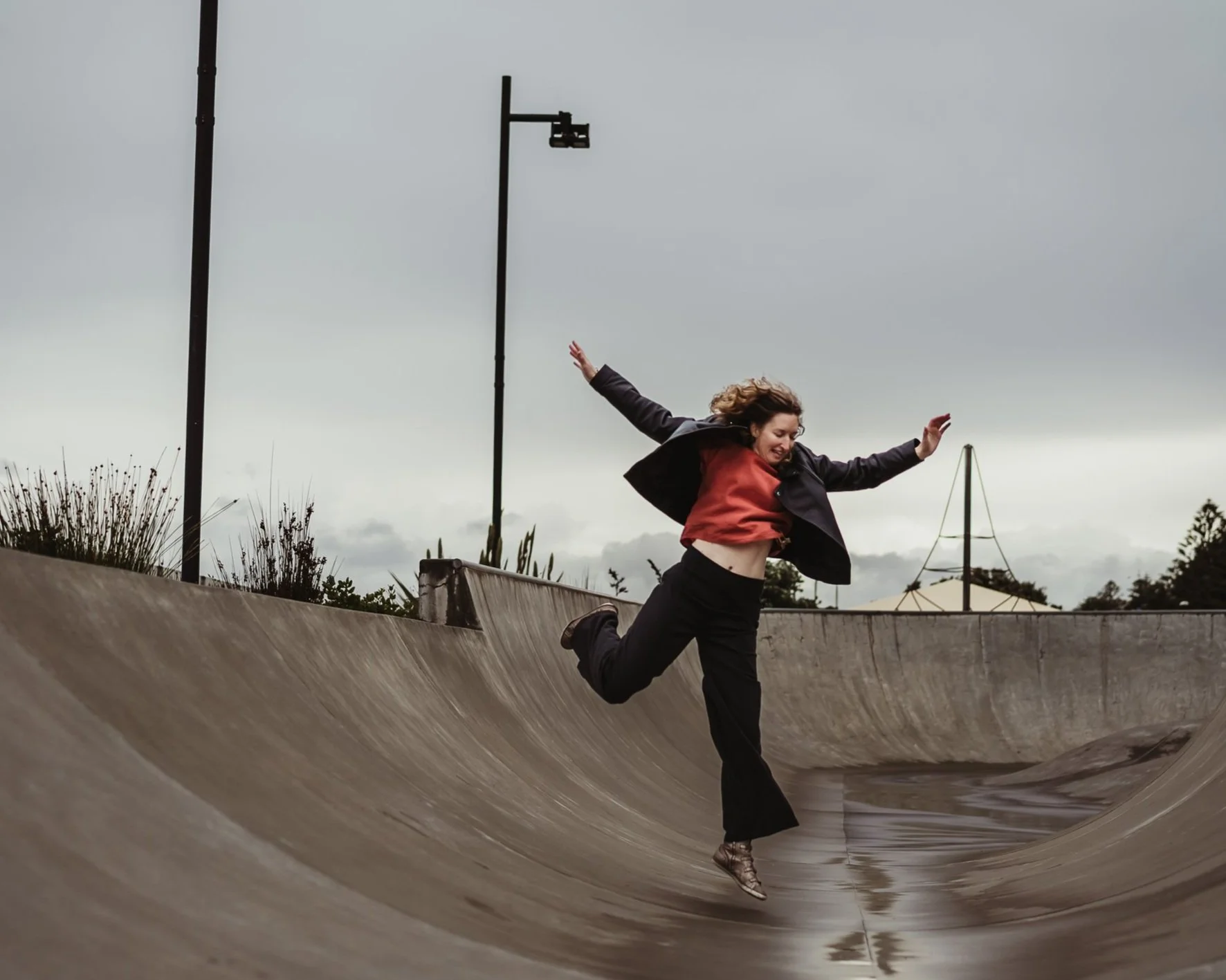 A woman is jumping in a skatepark bowl, balancing on one leg with arms outstretched on an overcast day.