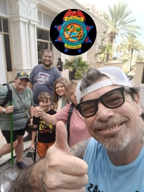 A group of six people smiling and giving thumbs up in front of a building with palm trees. There is a logo for a detective and metal detecting service above them.