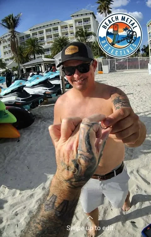 A man smiling on a beach holding hands with another person whose hand is tattooed, with Jet Skis parked nearby and a large hotel in the background, along with a