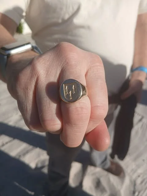 Close-up of a person's hand wearing a gold ring with a design, pointing toward the camera with a blurred background.